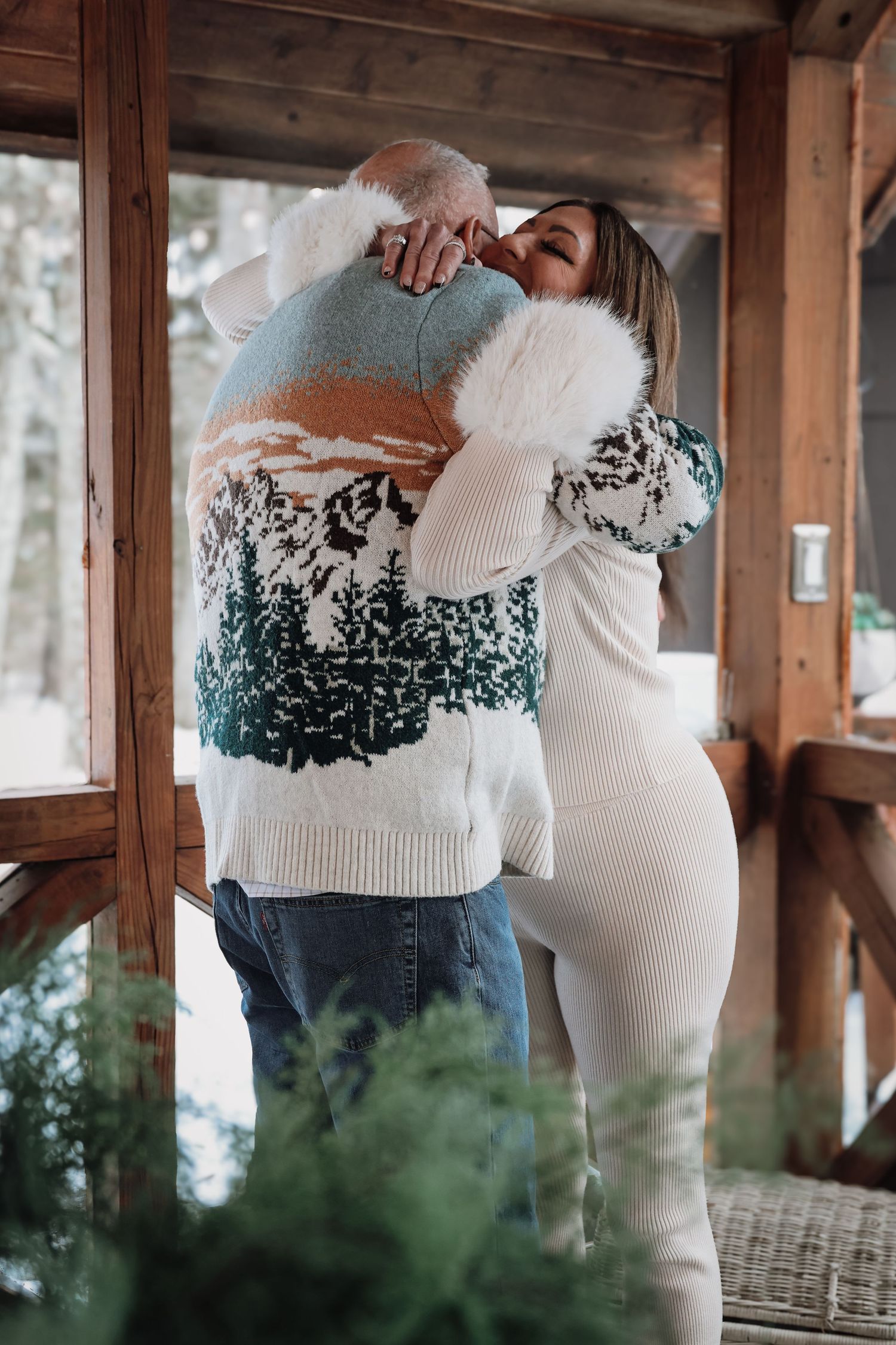 A couple embraces in a rustic wooden porch setting wearing matching white sweaters with green patterns.