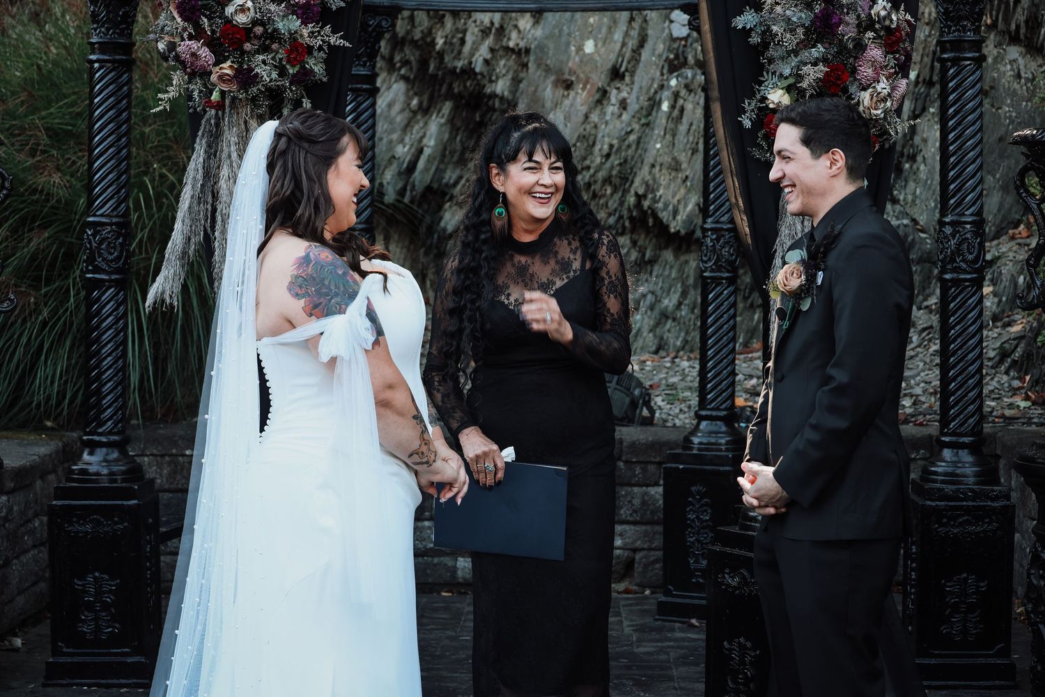 A wedding ceremony takes place outdoors with a bride in white dress and officiant under floral decorations.