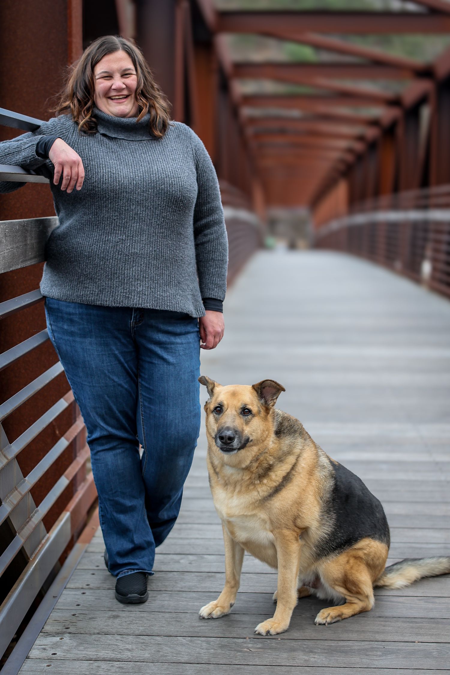 A German Shepherd mix dog sits loyally next to someone on a wooden deck on an overcast day.