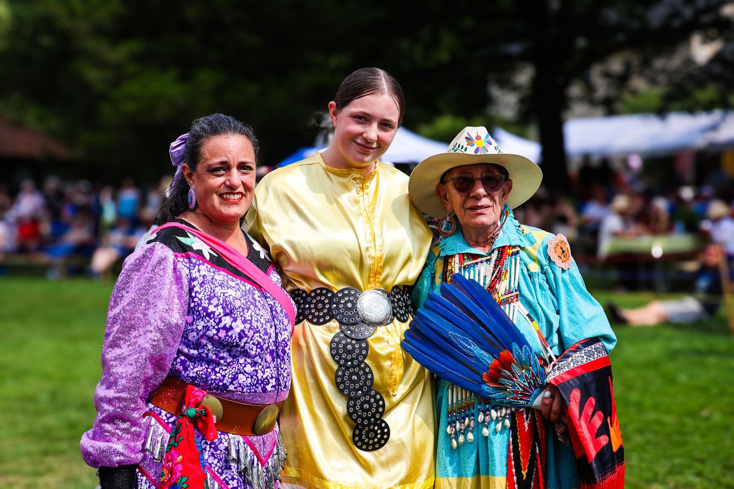 Colorfully dressed people in traditional Native American regalia gather at an outdoor powwow celebration.