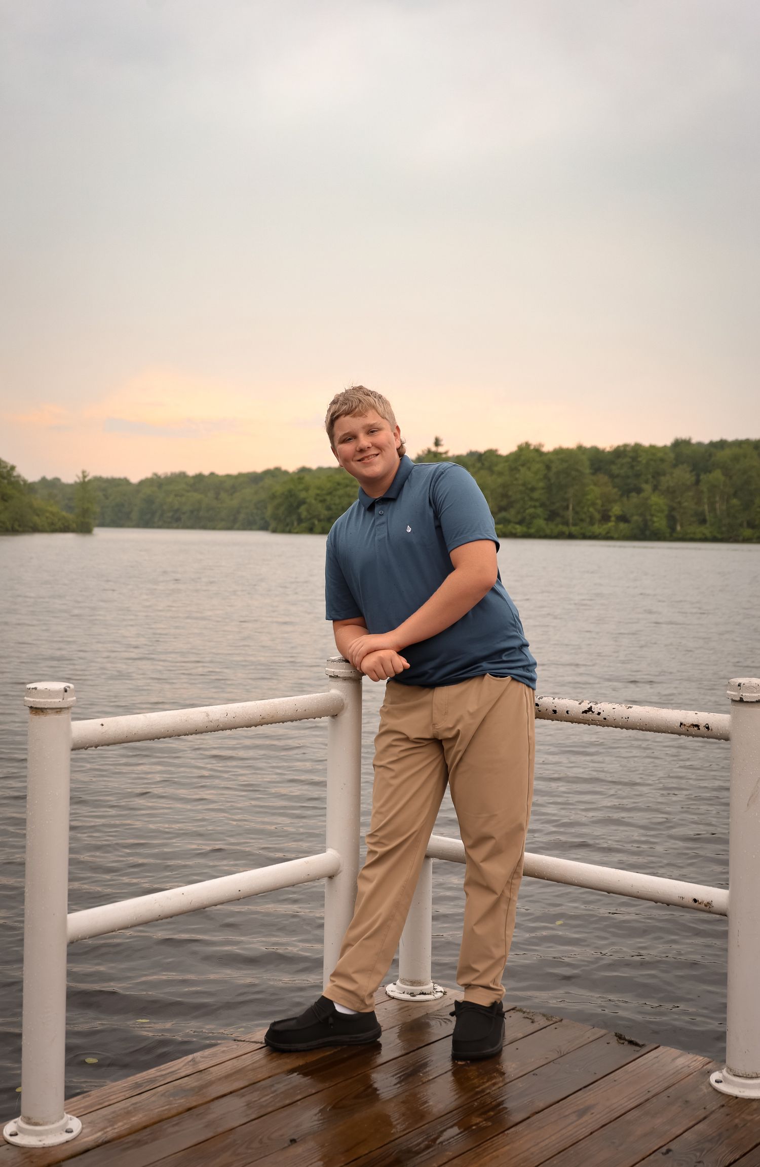 Person in blue shirt and khaki pants standing on wooden dock overlooking lake at sunset.
