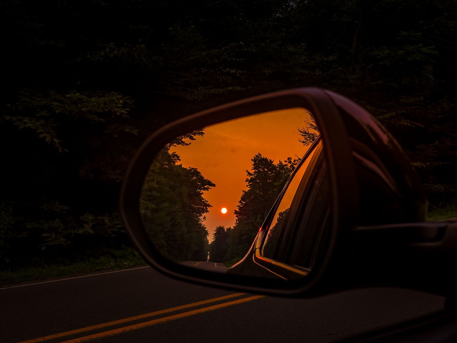 Orange sunset sky reflects in car side mirror against dark silhouette of trees and vehicle.