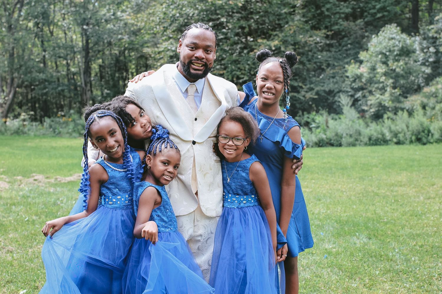 A group wearing coordinated blue dresses and a light colored suit pose together on a grassy lawn with trees in background.