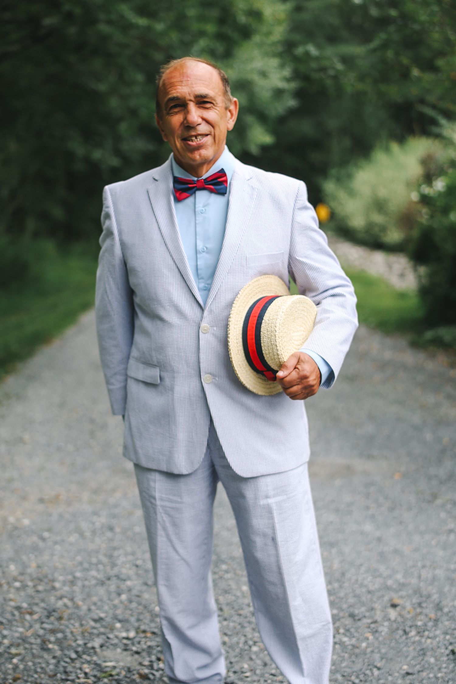A person in a light gray suit and red bowtie poses outdoors while holding a straw boater hat.