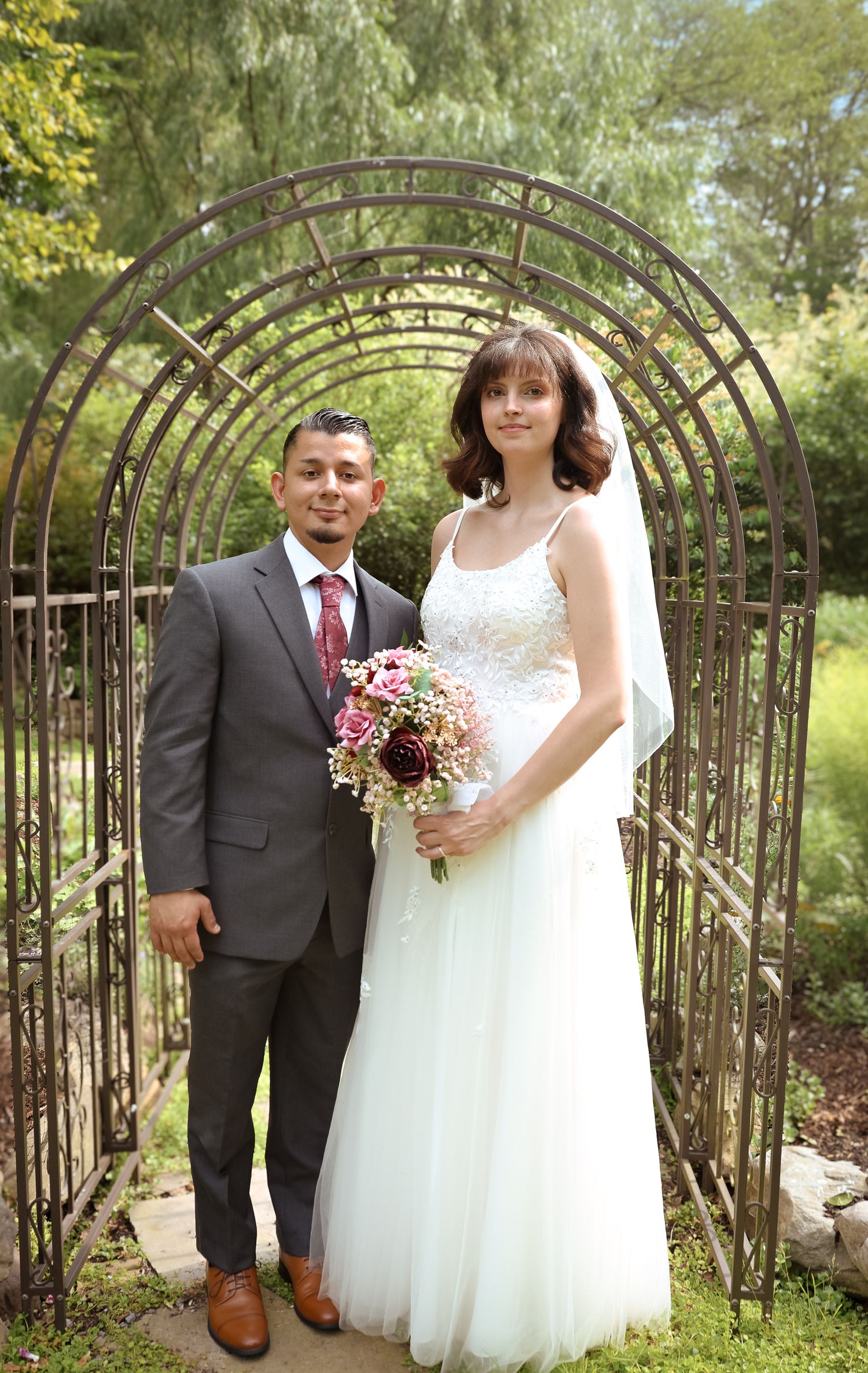 A couple poses in front of a beautiful garden archway during their outdoor wedding celebration.