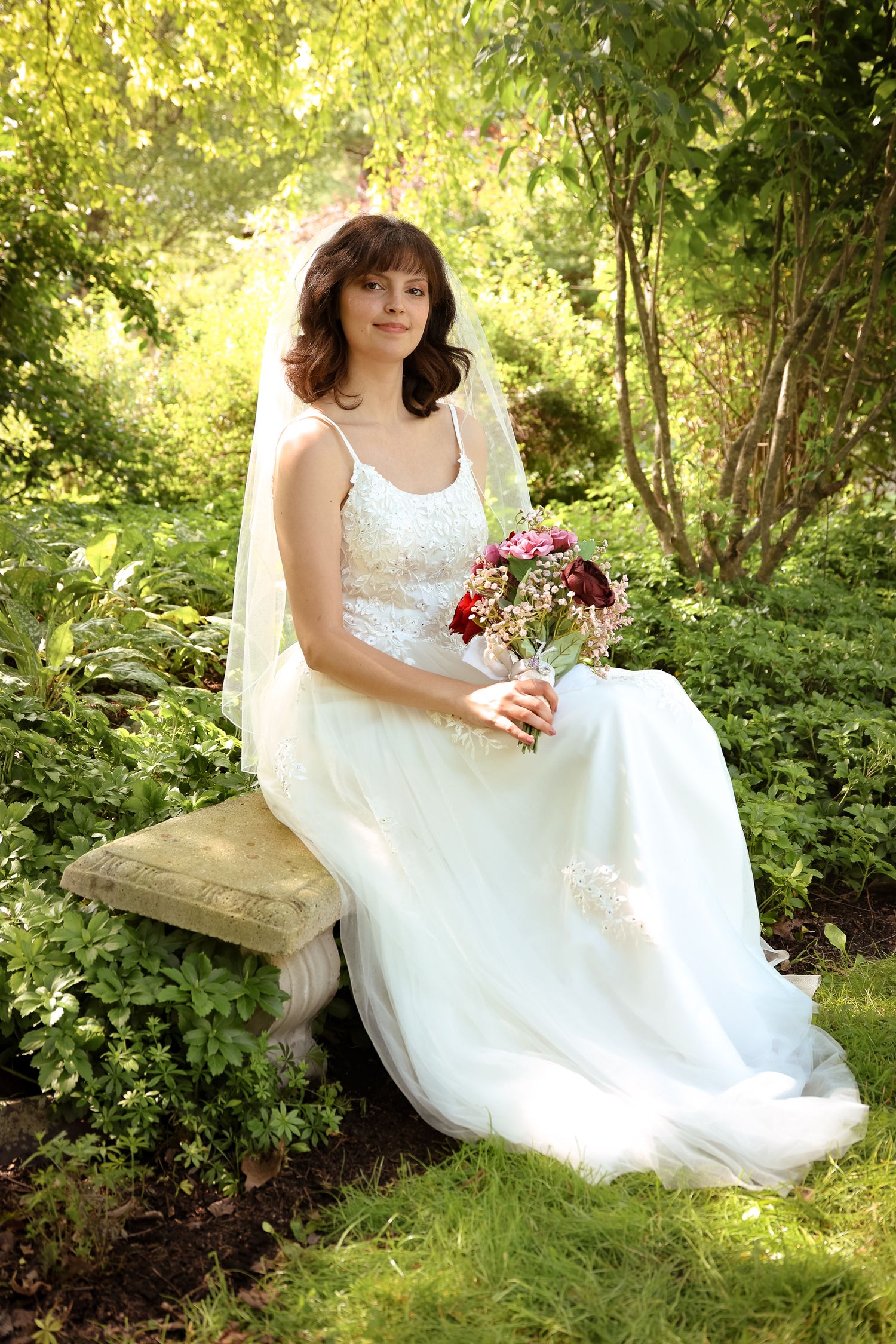 A person in a white wedding dress poses gracefully on a stone wall surrounded by lush summer greenery and flowers.