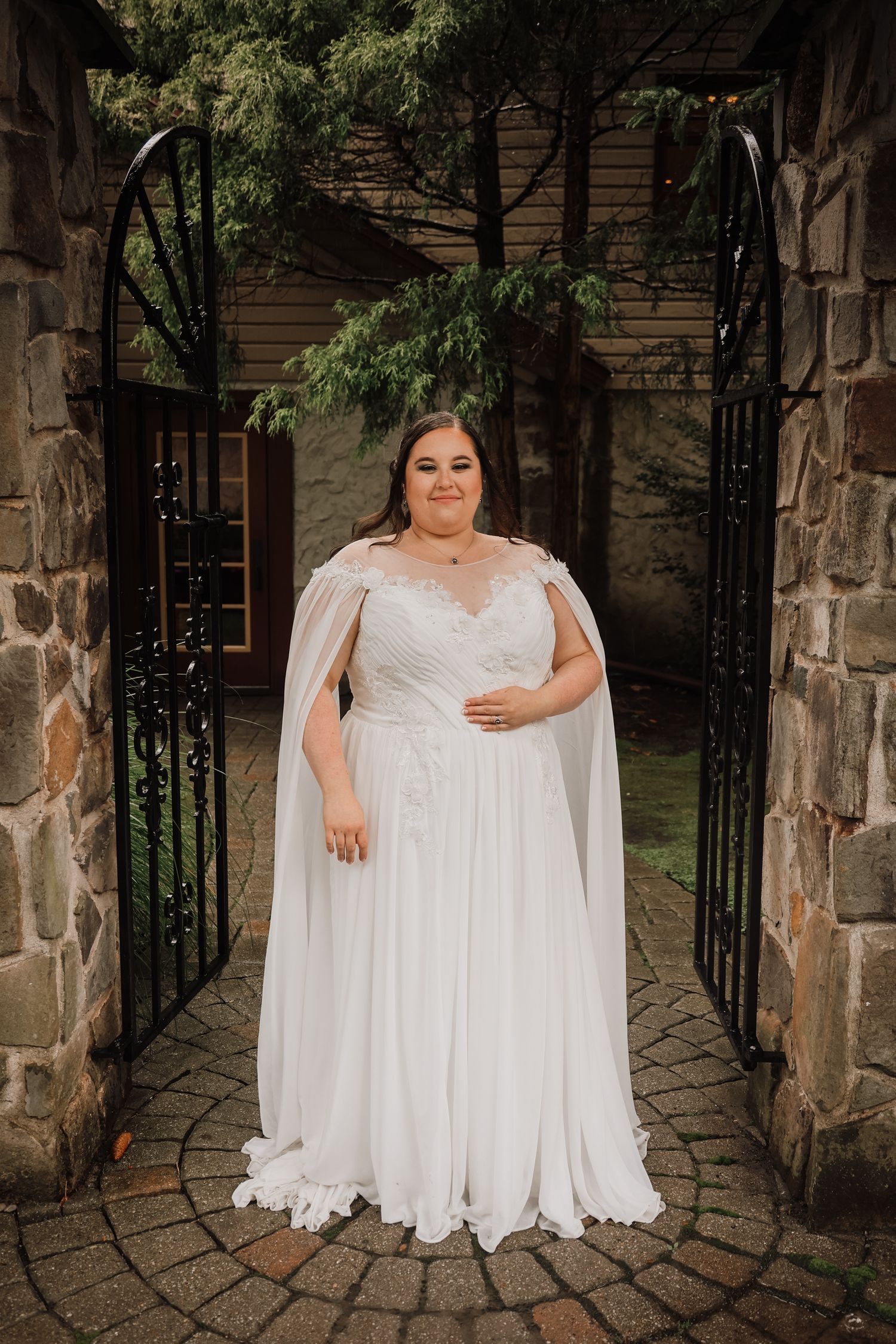 A bride in a flowing white cape dress stands between ornate stone pillars and iron gates in a garden setting.