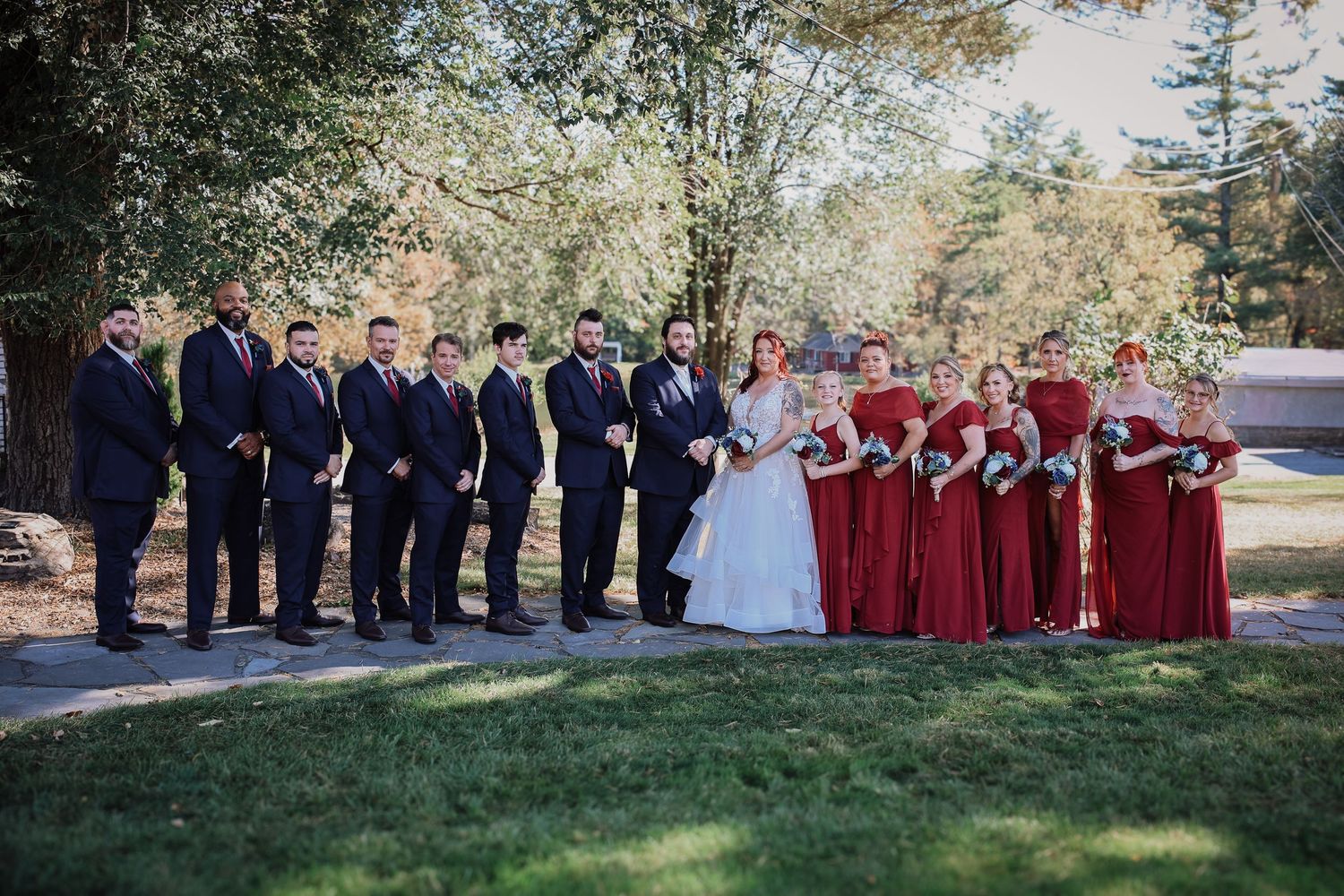 A wedding party stands in line outdoors with bridesmaids in burgundy dresses and groomsmen in navy suits.
