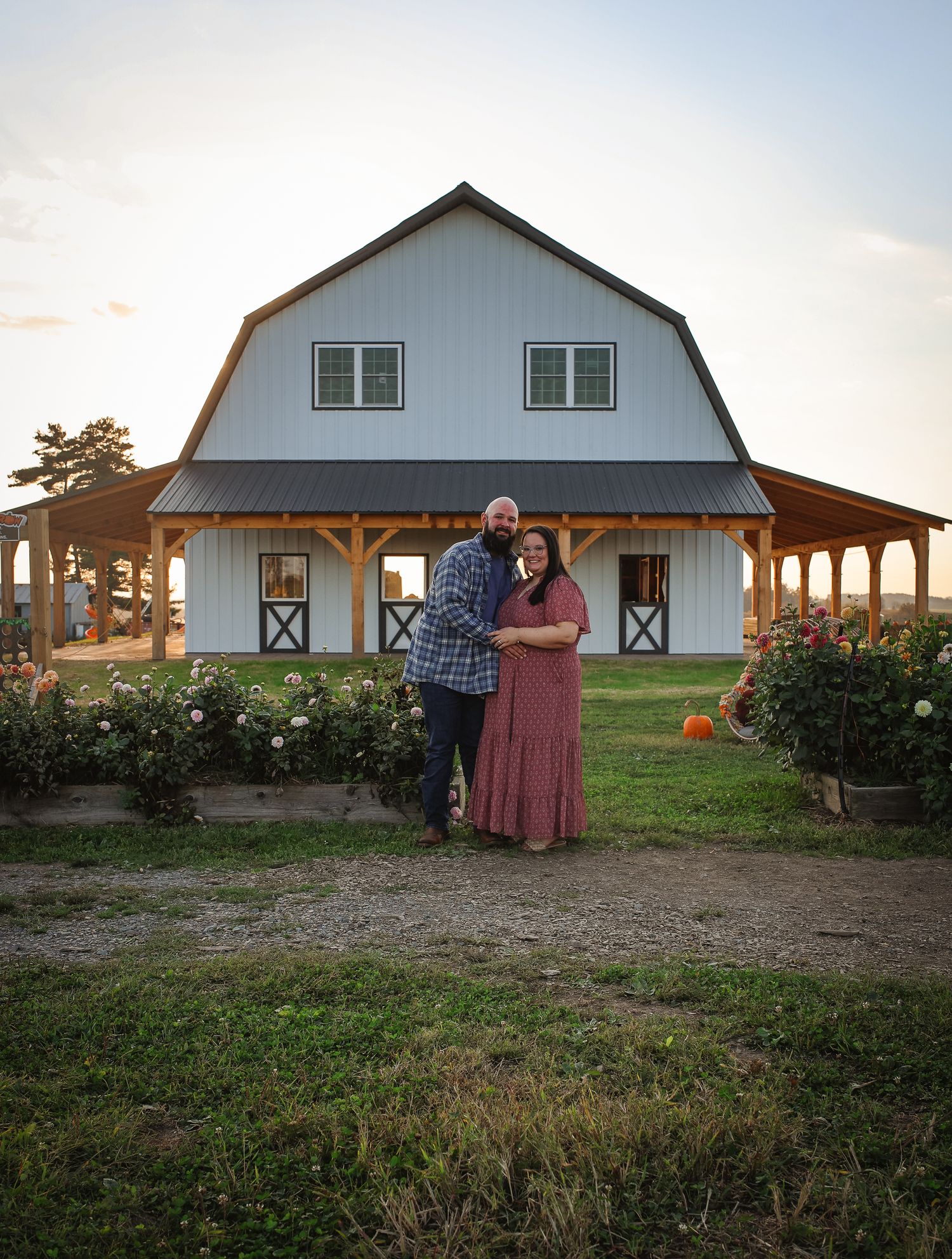 A couple poses together at sunset in front of a white barn with a covered porch and rose garden.