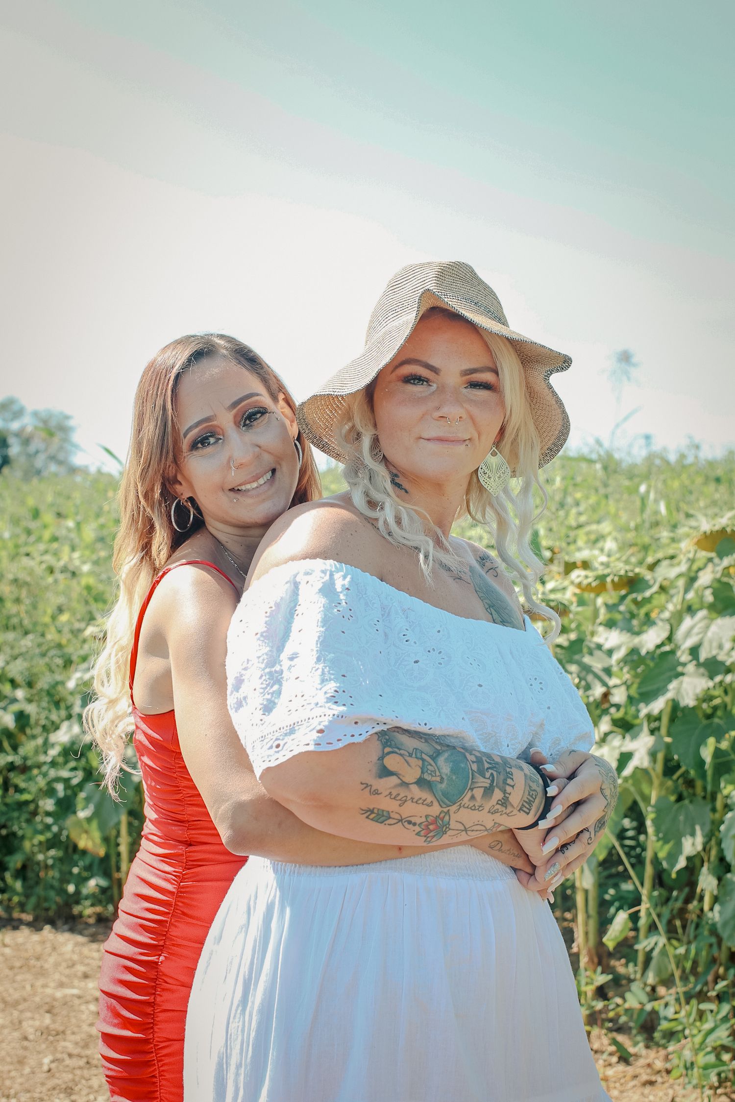 Two people embrace in a sunlit field of sunflowers wearing red and white outfits.