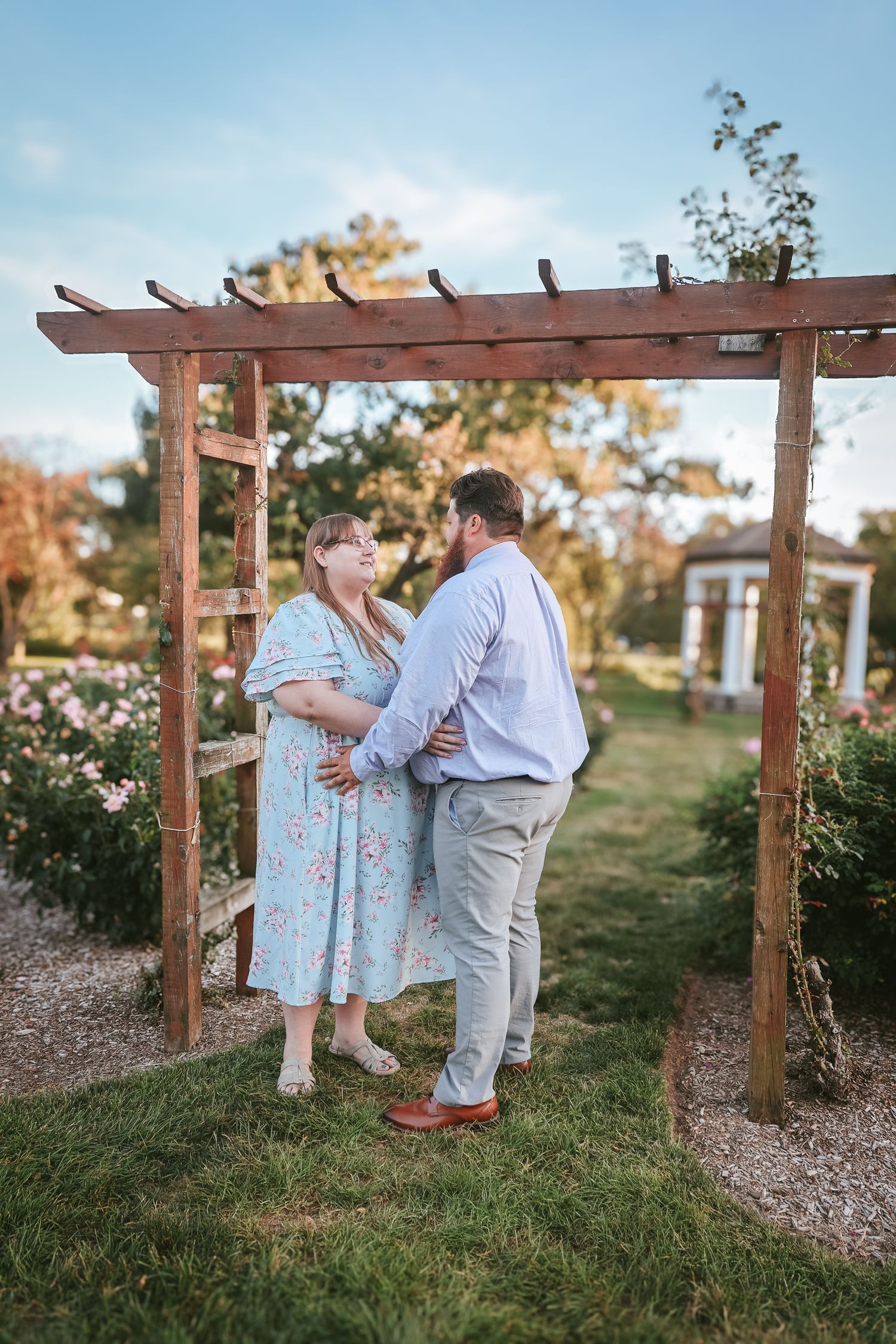 A couple in a garden setting embraces under a wooden pergola during sunset.