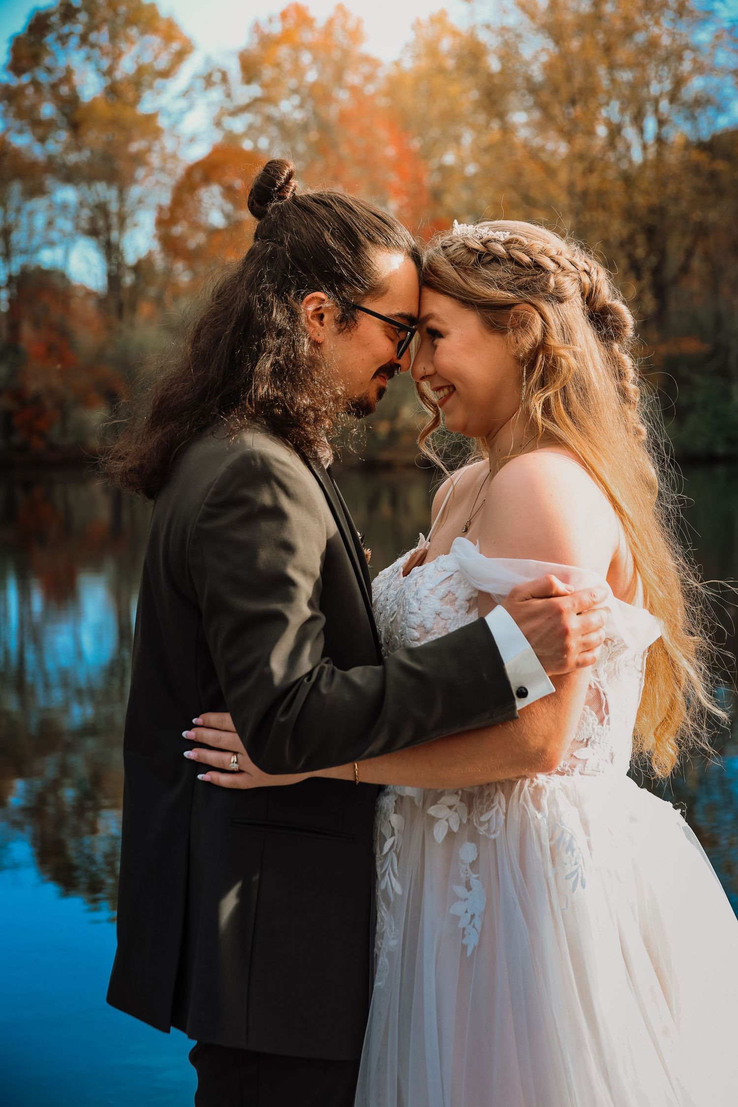 A romantic couple embraces by a lake during autumn, with the bride in white and groom in dark suit against fall foliage.
