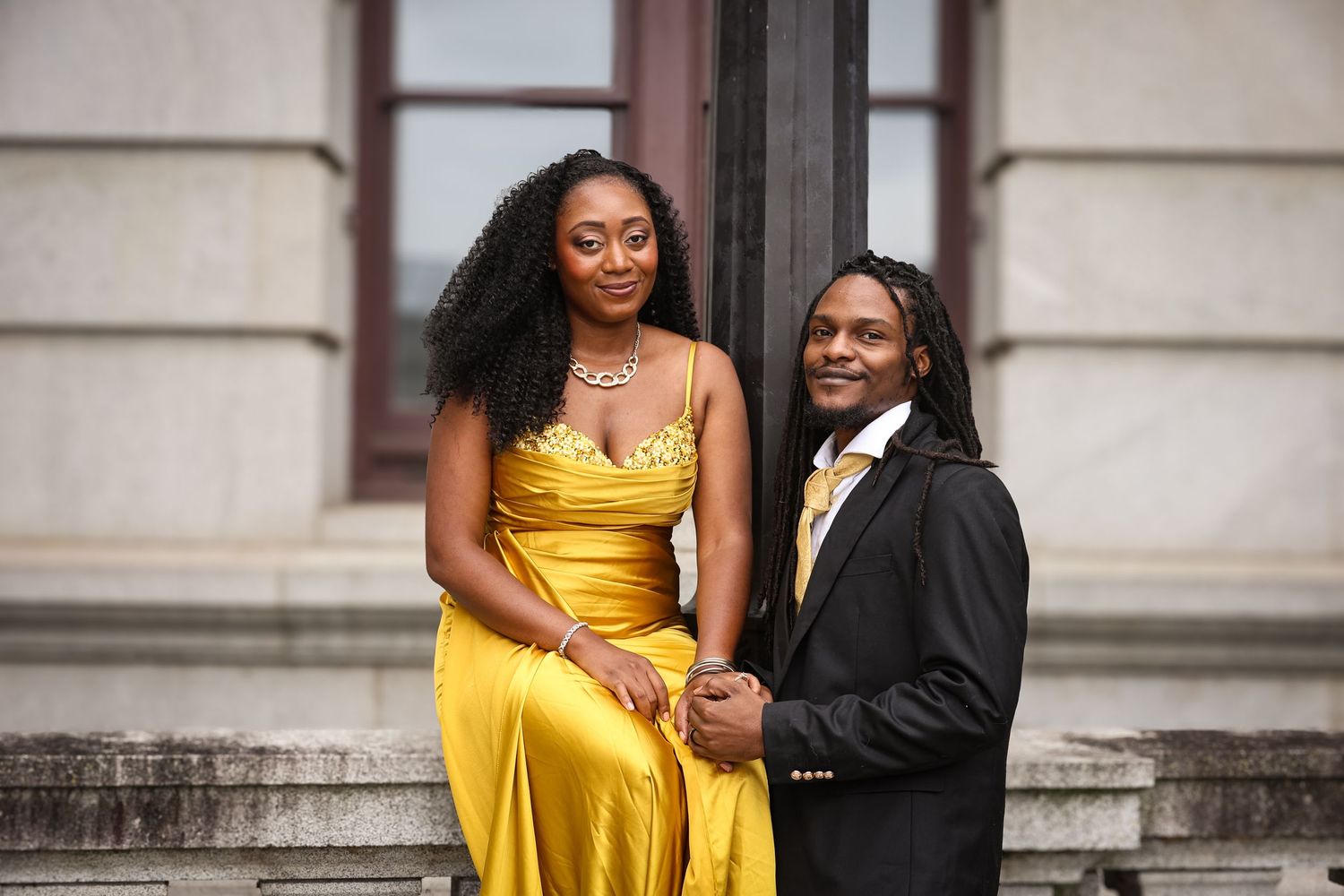 A couple in formal attire, with a yellow dress and black suit, pose together for photos on stone steps.