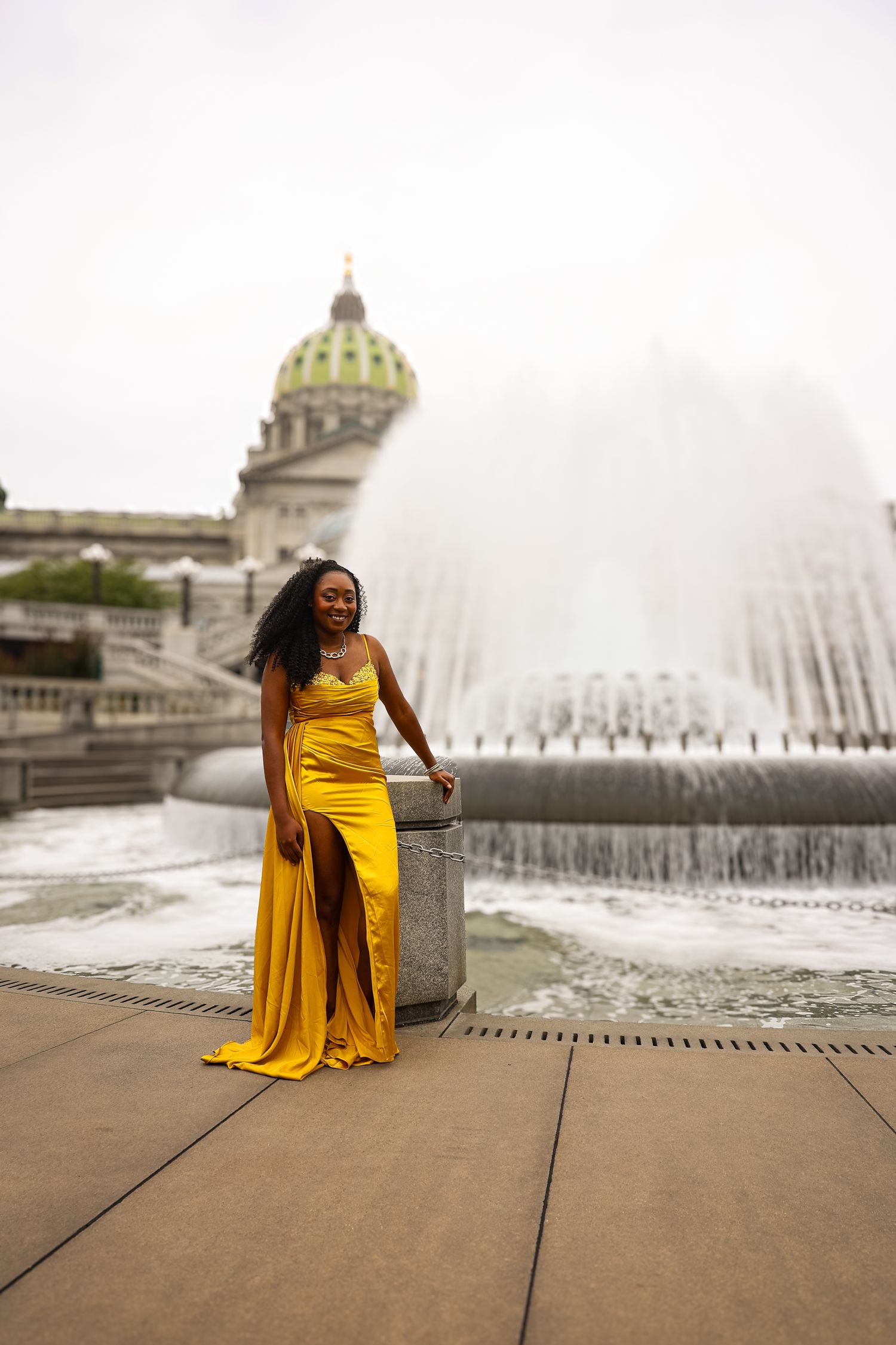 Elegant yellow evening gown photographed in front of a grand domed building and fountain on a cloudy day.