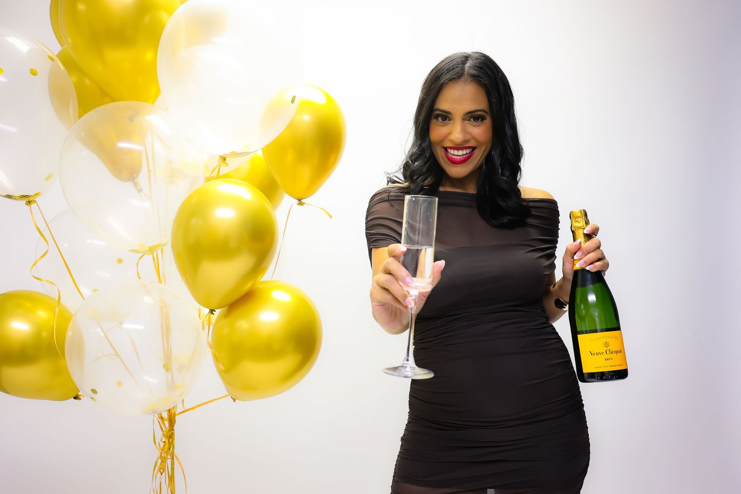 Woman in black dress holding champagne glass and bottle with gold and white balloons celebrating a festive occasion.