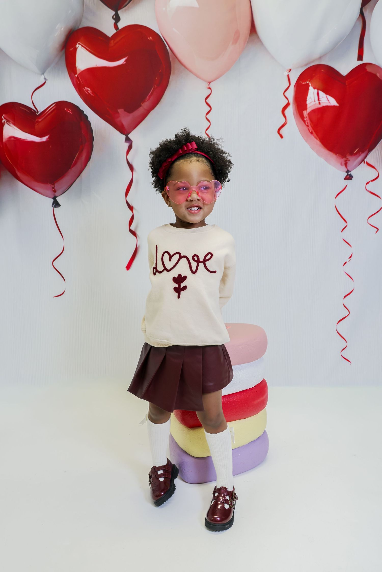 A young girl in a love sweatshirt and skirt poses with heart balloons for Valentine's Day.