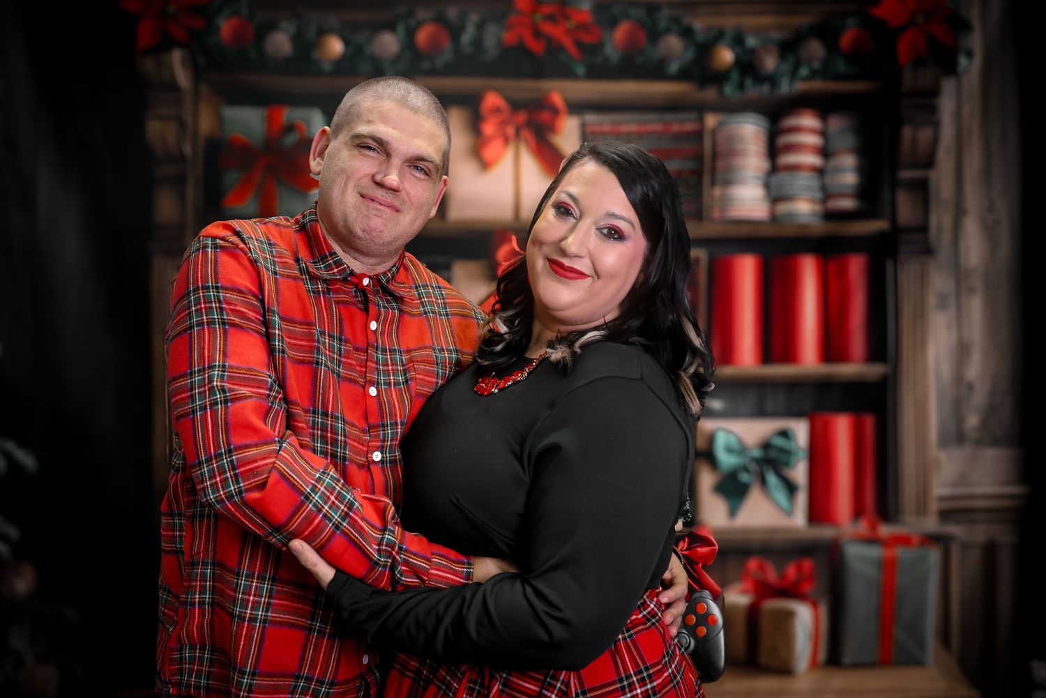 Couple posing together in matching red plaid outfits against a festive Christmas backdrop with gifts and decorations.
