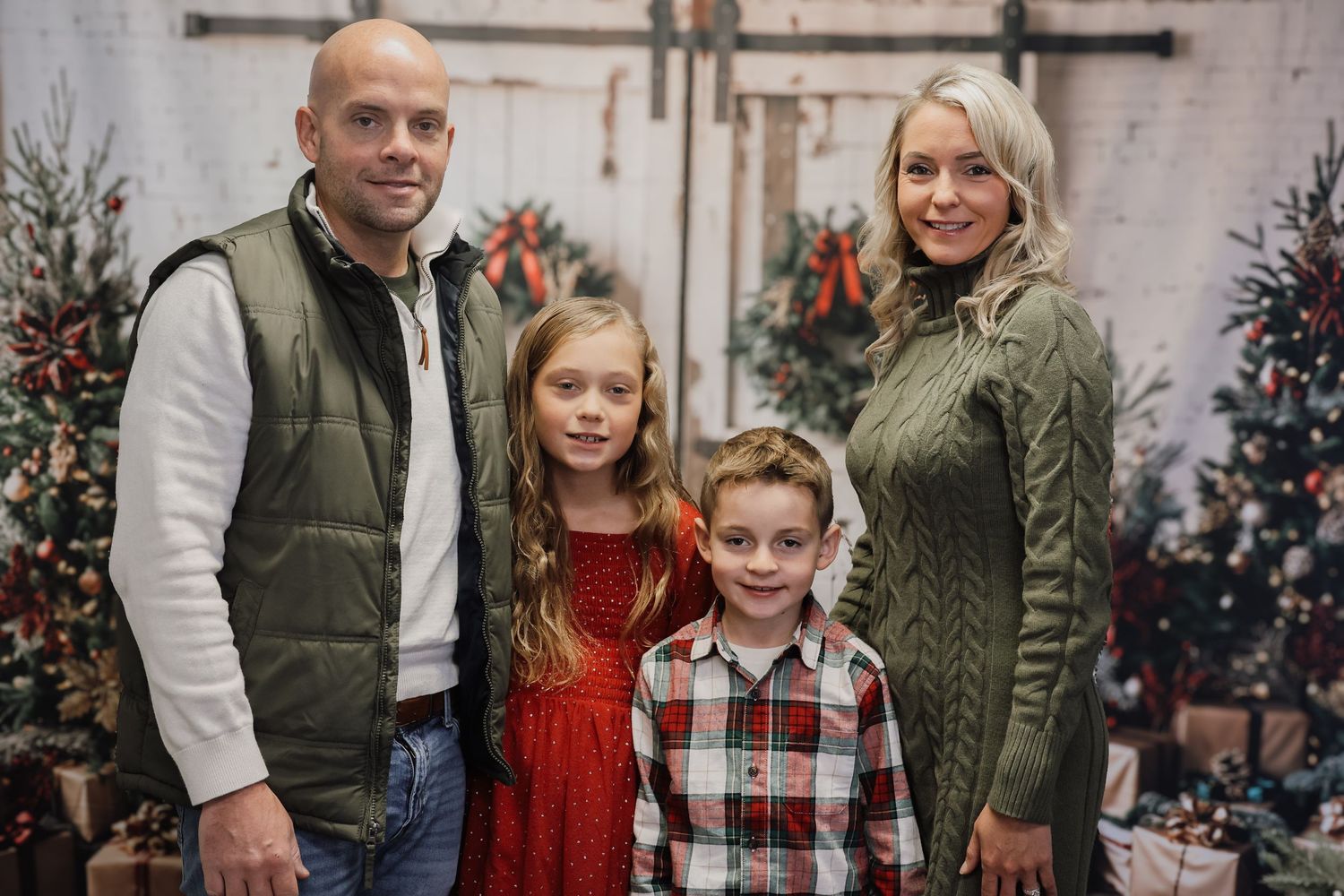 Family of four posing for a Christmas portrait with festive holiday decorations and Christmas trees in the background.