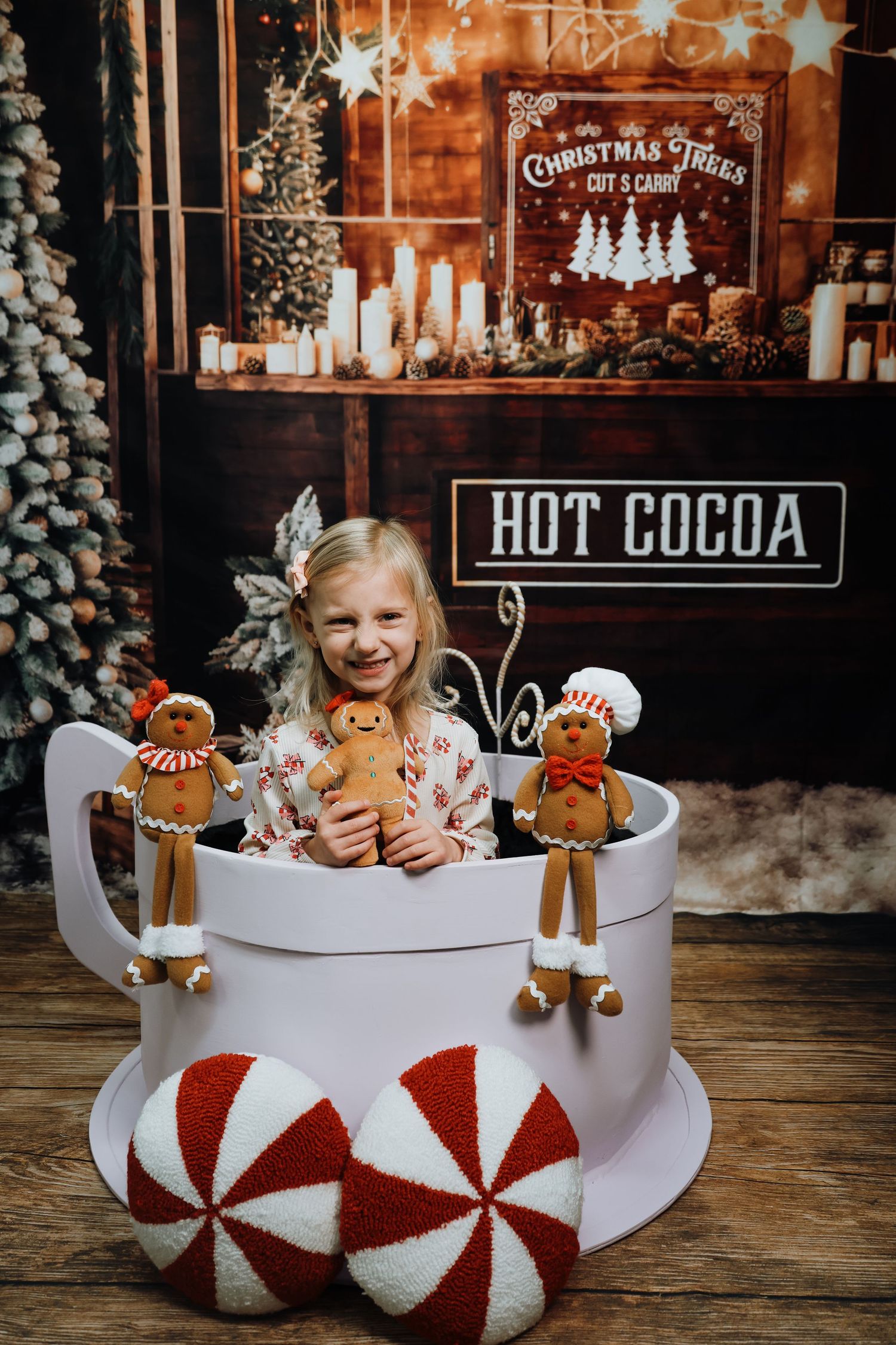 Young girl sitting in giant hot cocoa mug prop with gingerbread decorations in festive Christmas photo studio setting.
