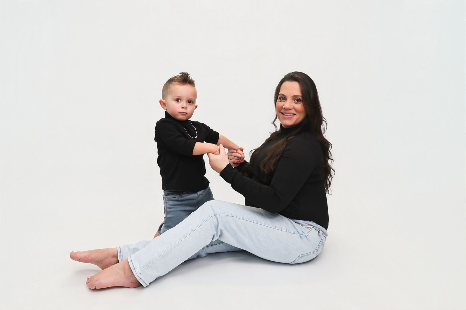 A smiling woman in black top and jeans sits cross-legged with a standing toddler in matching outfit on white background.