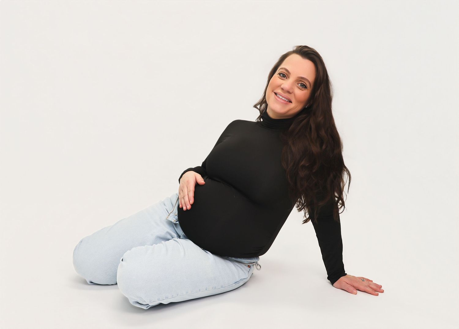 Smiling pregnant woman in black turtleneck and light jeans sitting on white background.