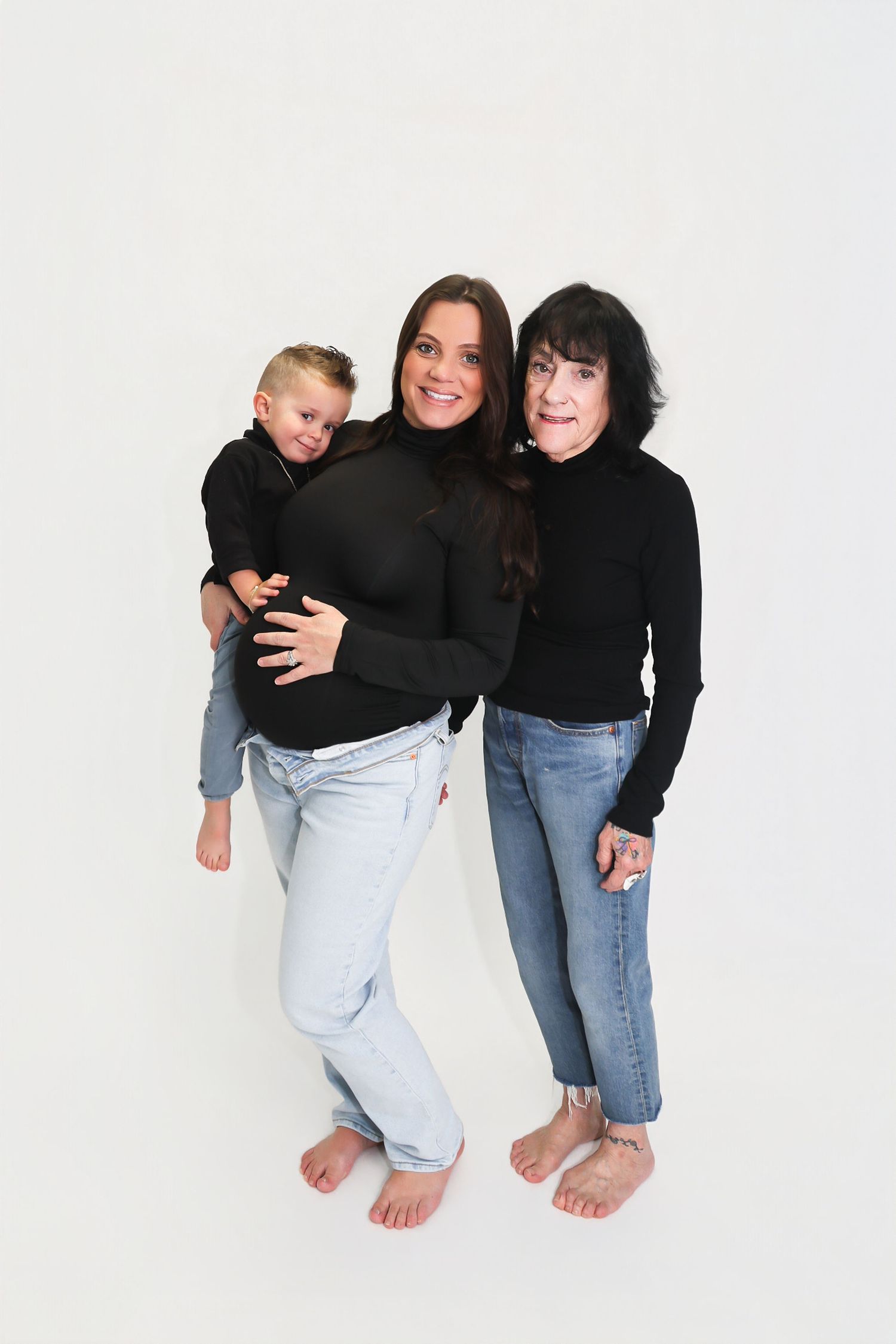 Three generations in matching black tops and jeans pose together barefoot against a white background.