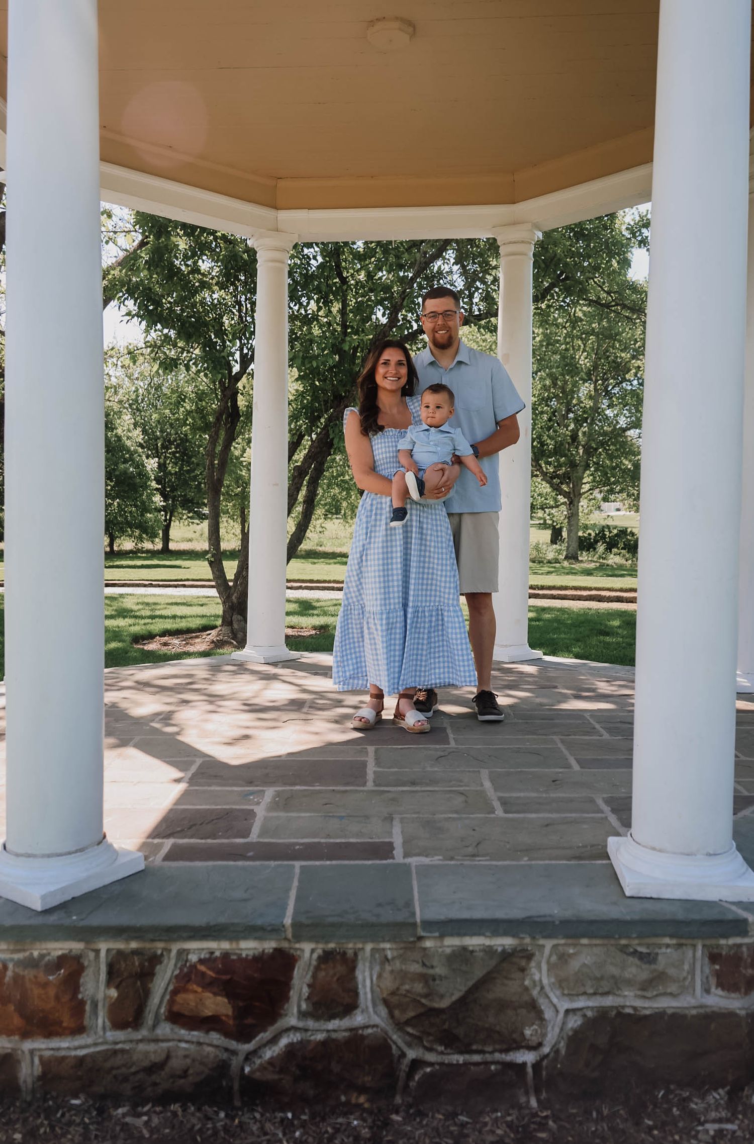 Family of three posing under a white gazebo in a lush park, wearing coordinated blue outfits.