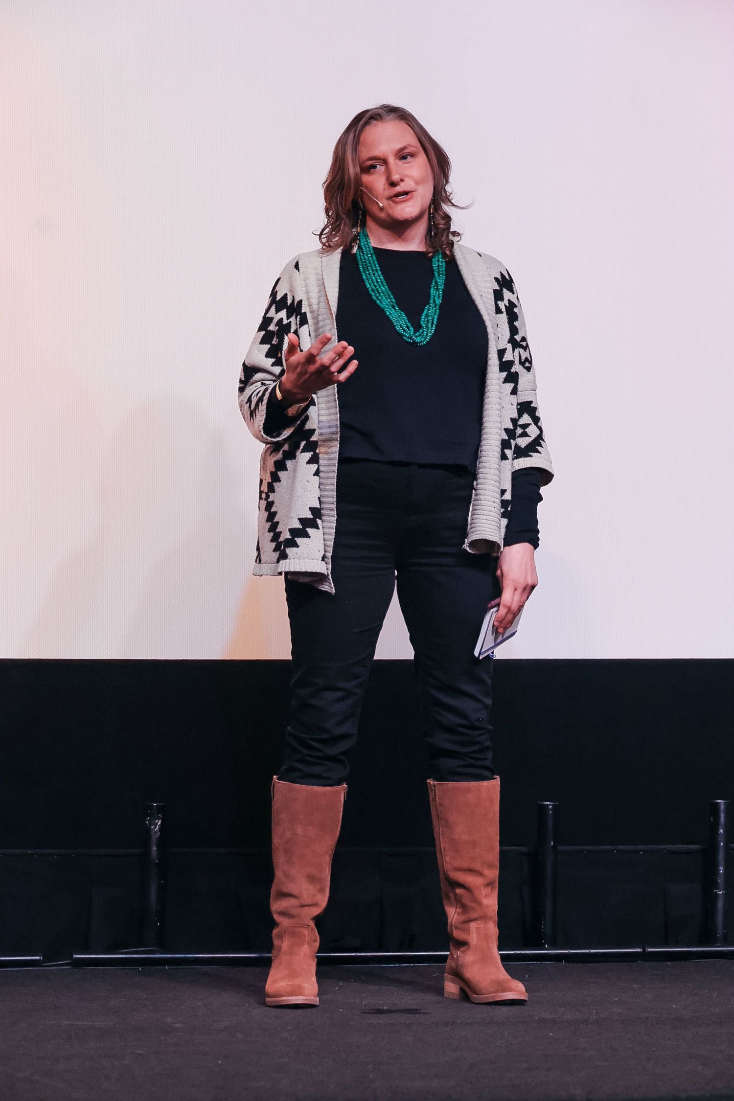 Woman in patterned cardigan, black outfit, turquoise necklace, and tan boots speaking on stage.