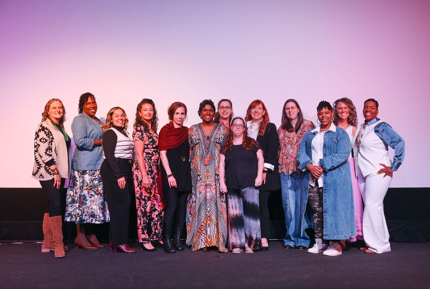 Diverse group of women posing together on stage in front of a pink and purple gradient backdrop.