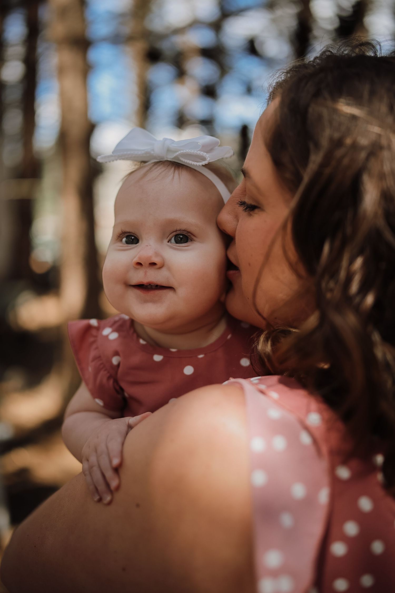 A mother kisses her smiling baby girl wearing a white bow headband and pink polka dot dress outdoors.