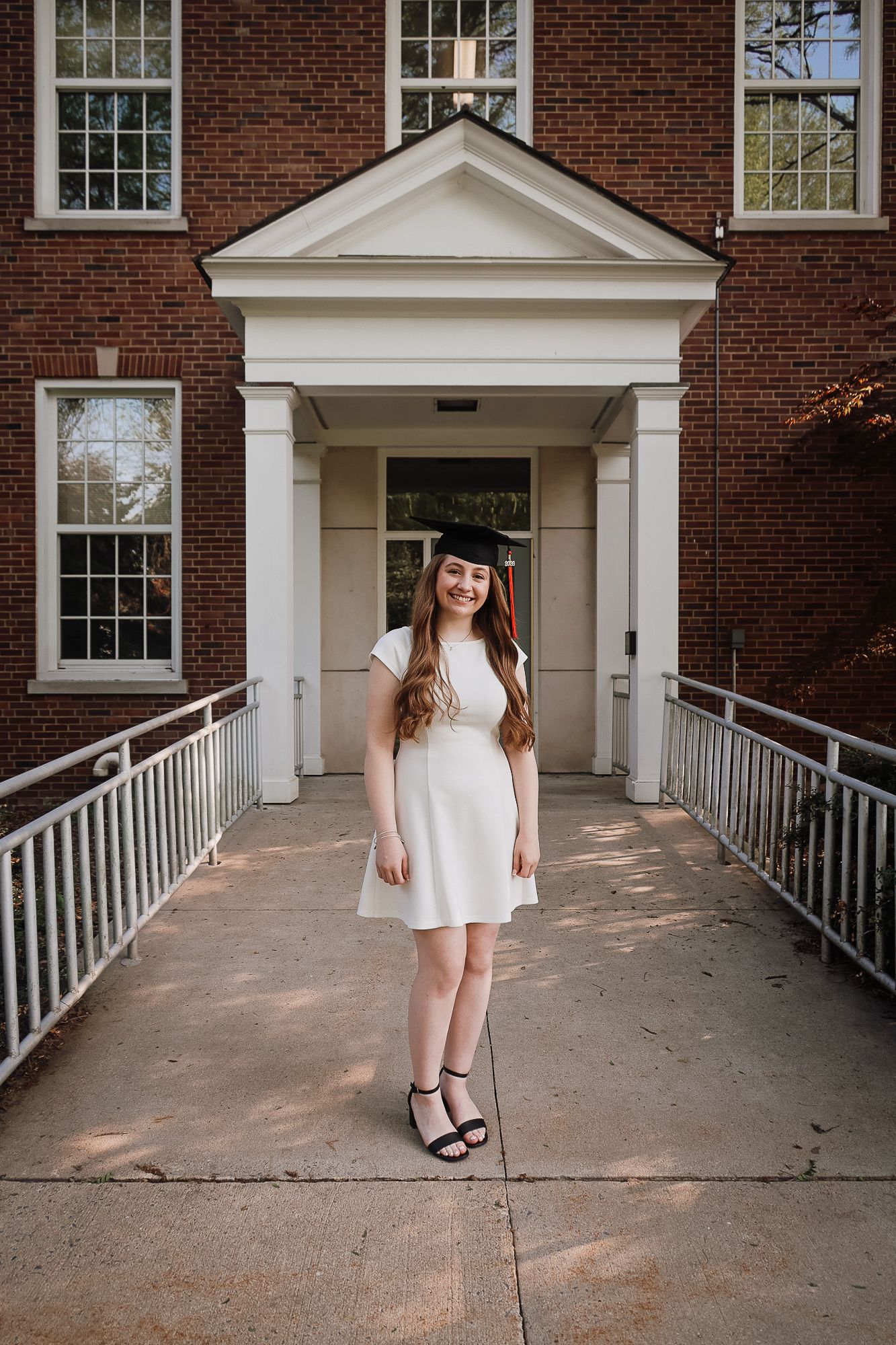 Young woman in white dress and graduation cap poses proudly on walkway in front of brick building.