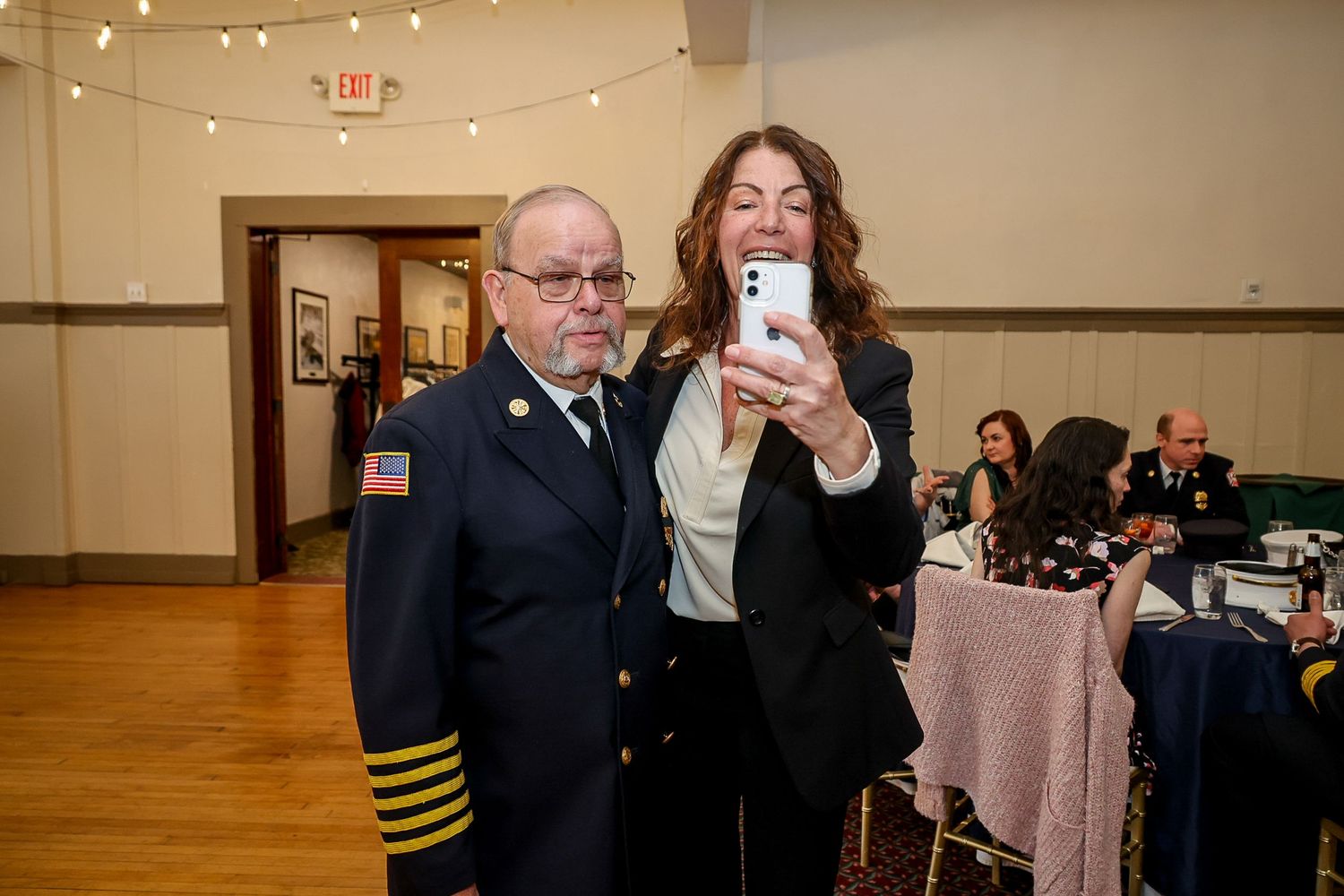 A woman takes a selfie with a man in a fire department uniform at a formal indoor event.