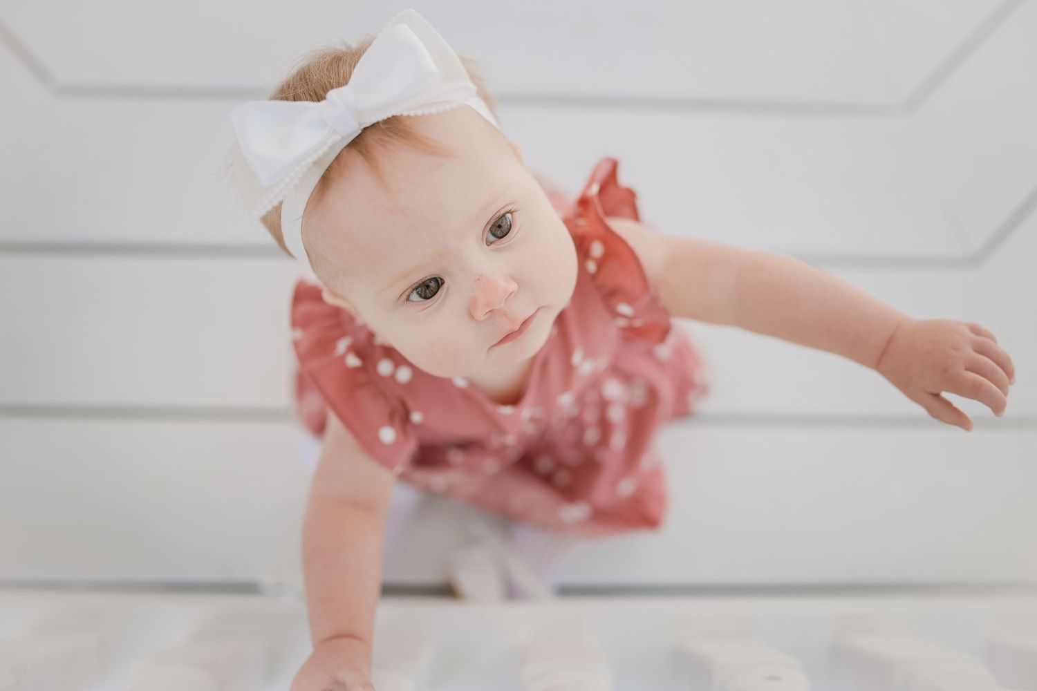 Adorable baby girl in pink polka dot outfit and white bow headband crawling on a white surface.