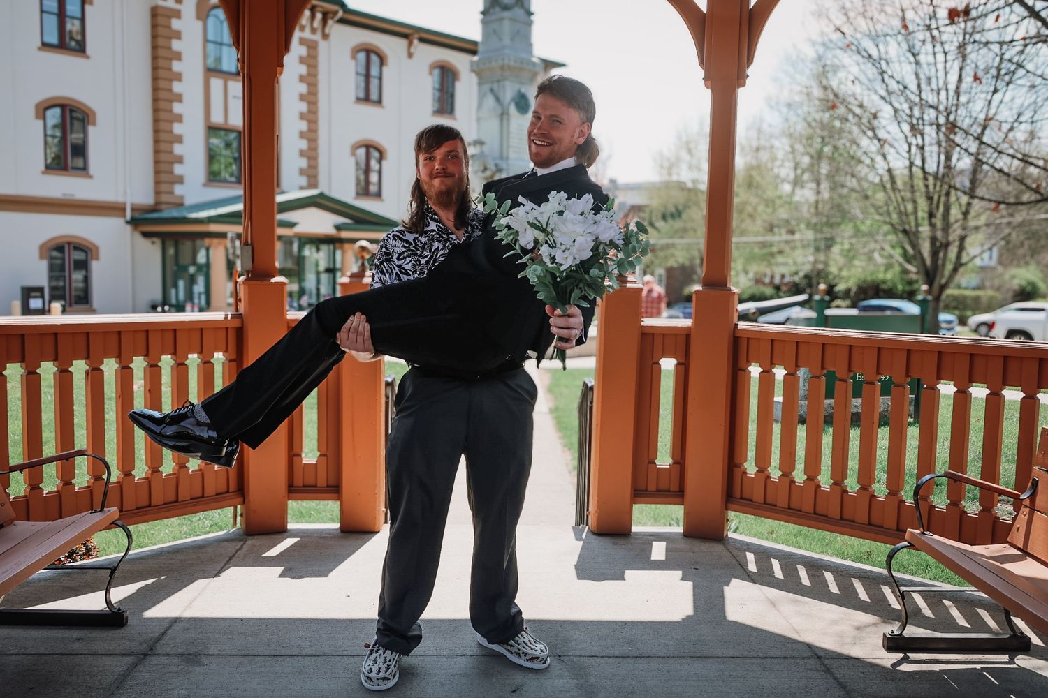 A groom lifts his bride in a gazebo, holding white flowers, with a charming building in the background on a sunny day.