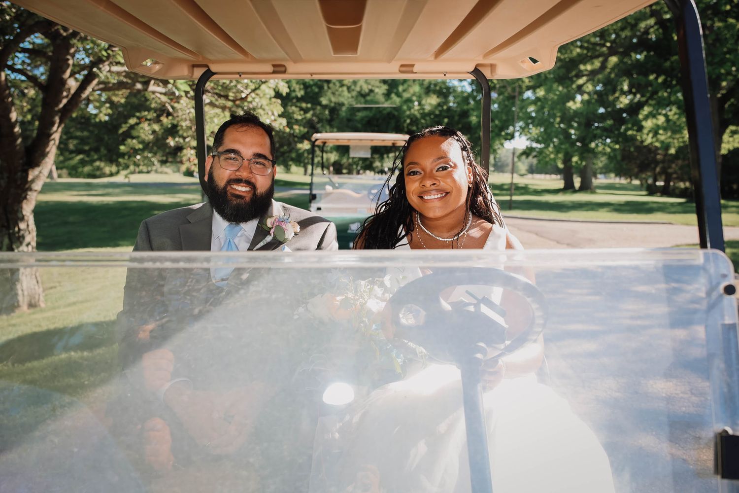 Bride and groom smiling together in a golf cart outdoors on a sunny wedding day.