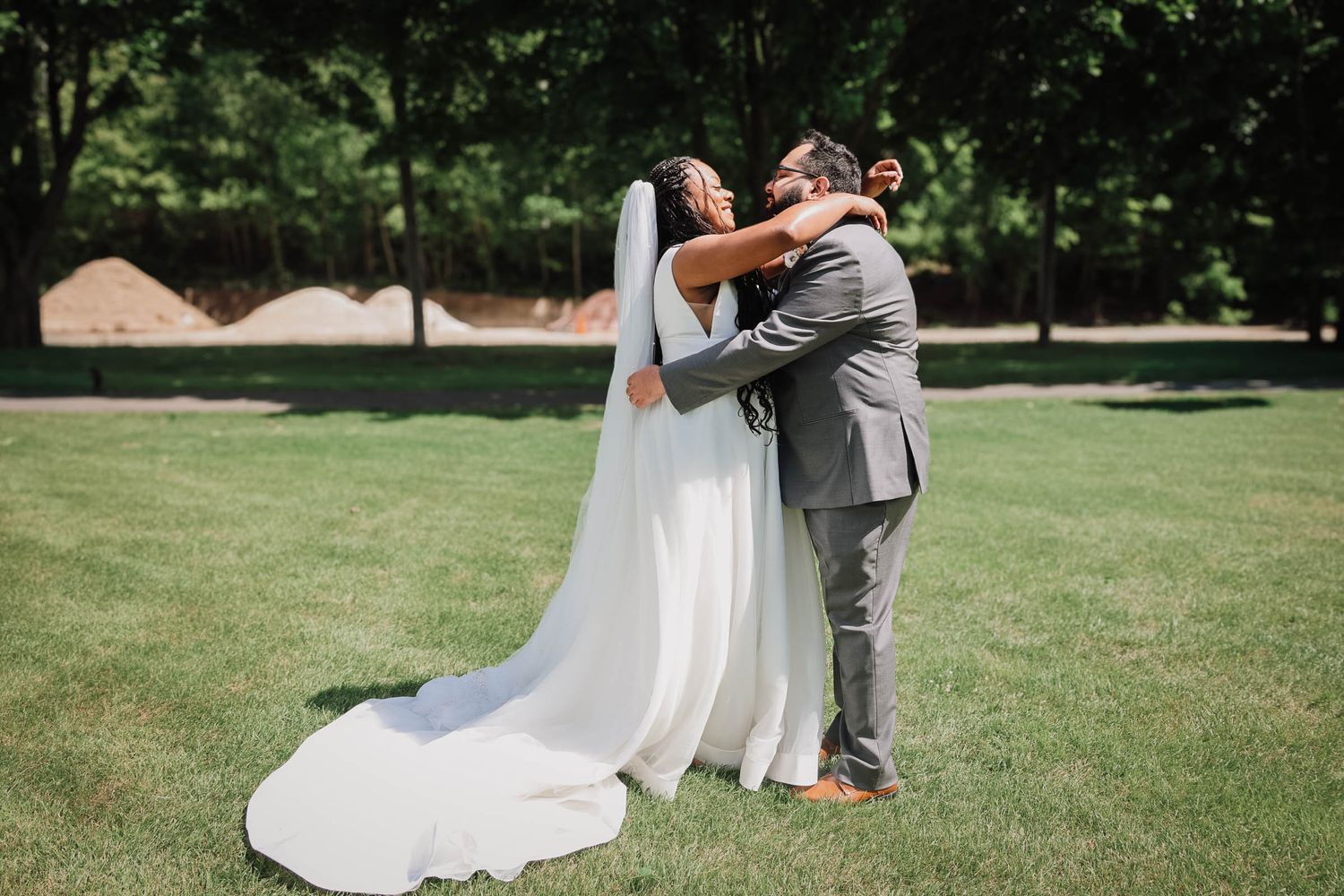 Bride and groom share a heartfelt embrace on green lawn, bride wearing flowing white gown with long veil.