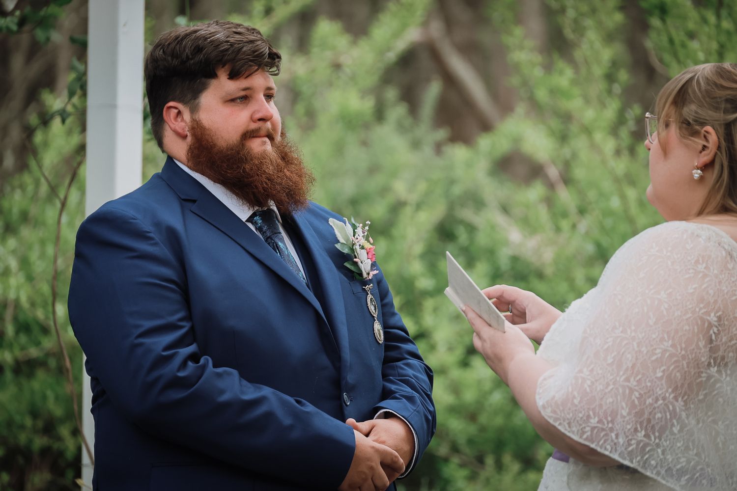 Groom in blue suit with beard listens as bride reads wedding vows during outdoor ceremony.