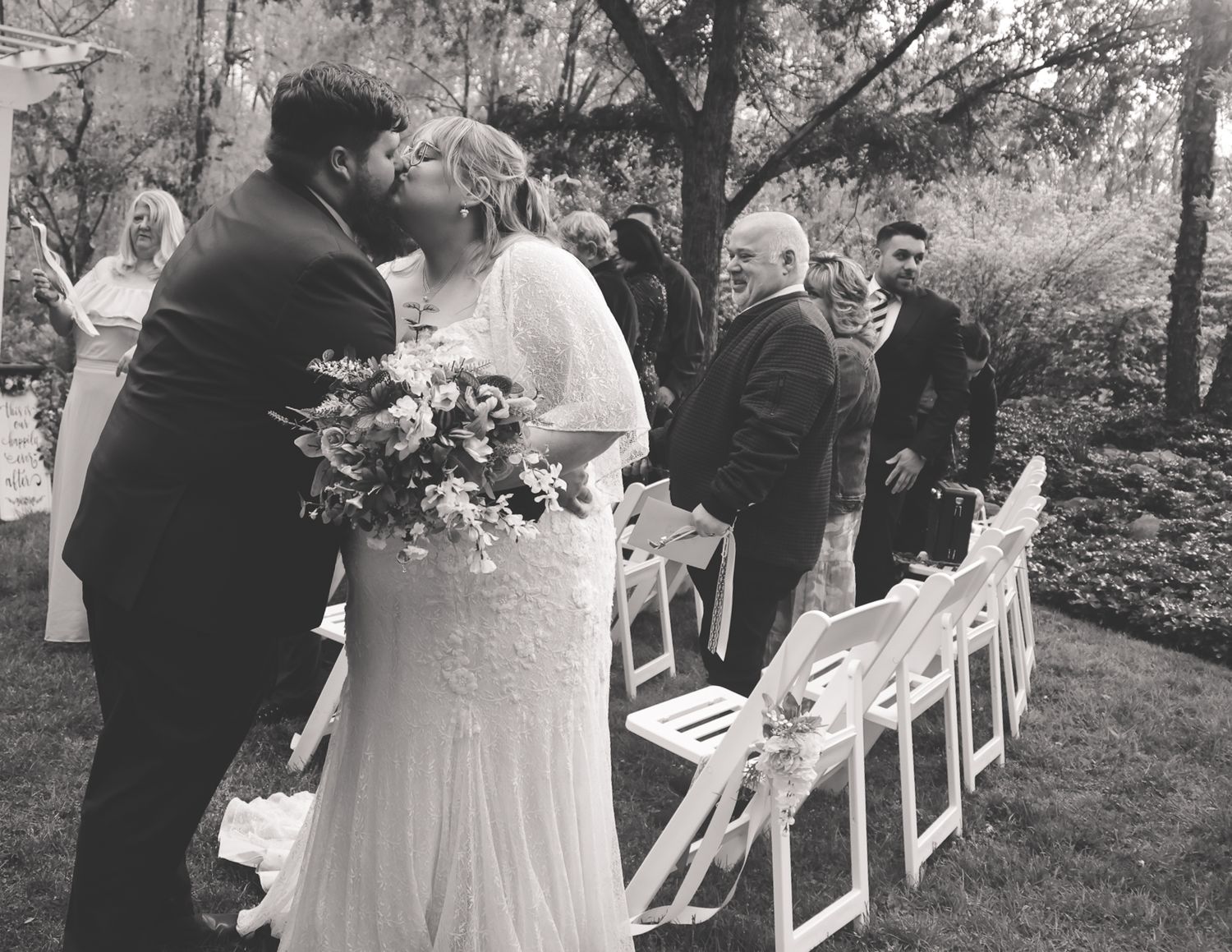 Black and white outdoor wedding ceremony photo of bride and groom kissing, with white chairs and guests in background.