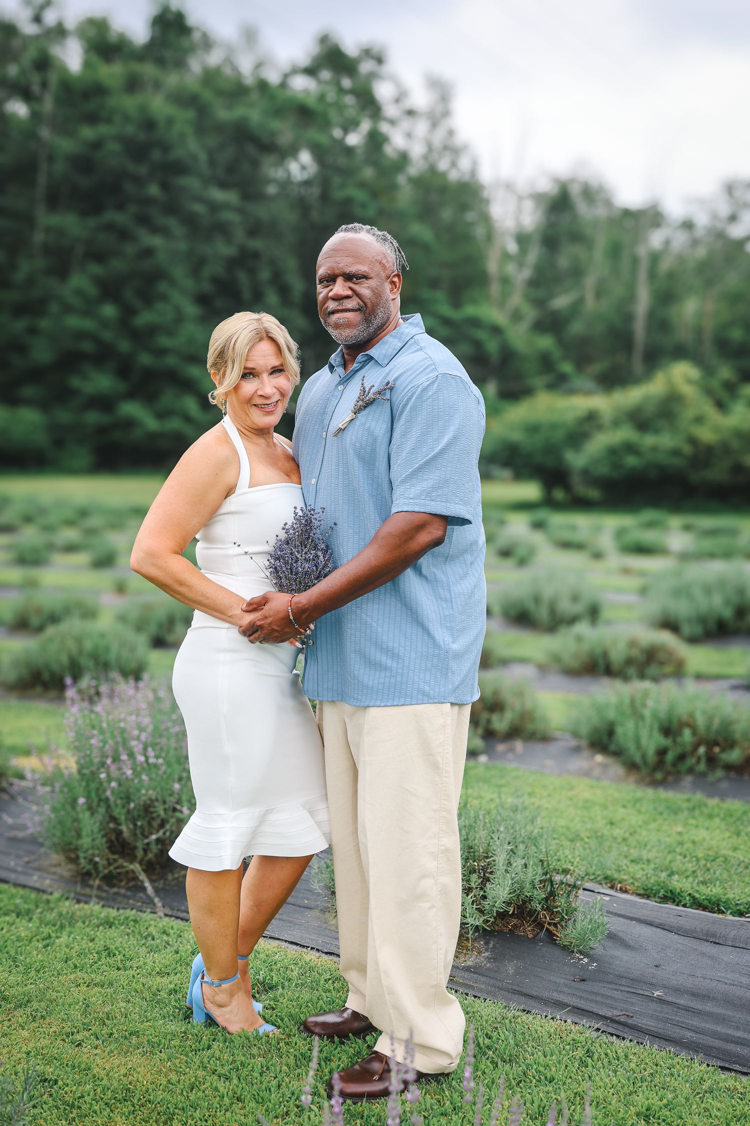 A couple poses together in a lavender garden wearing coordinating white and blue summer outfits.