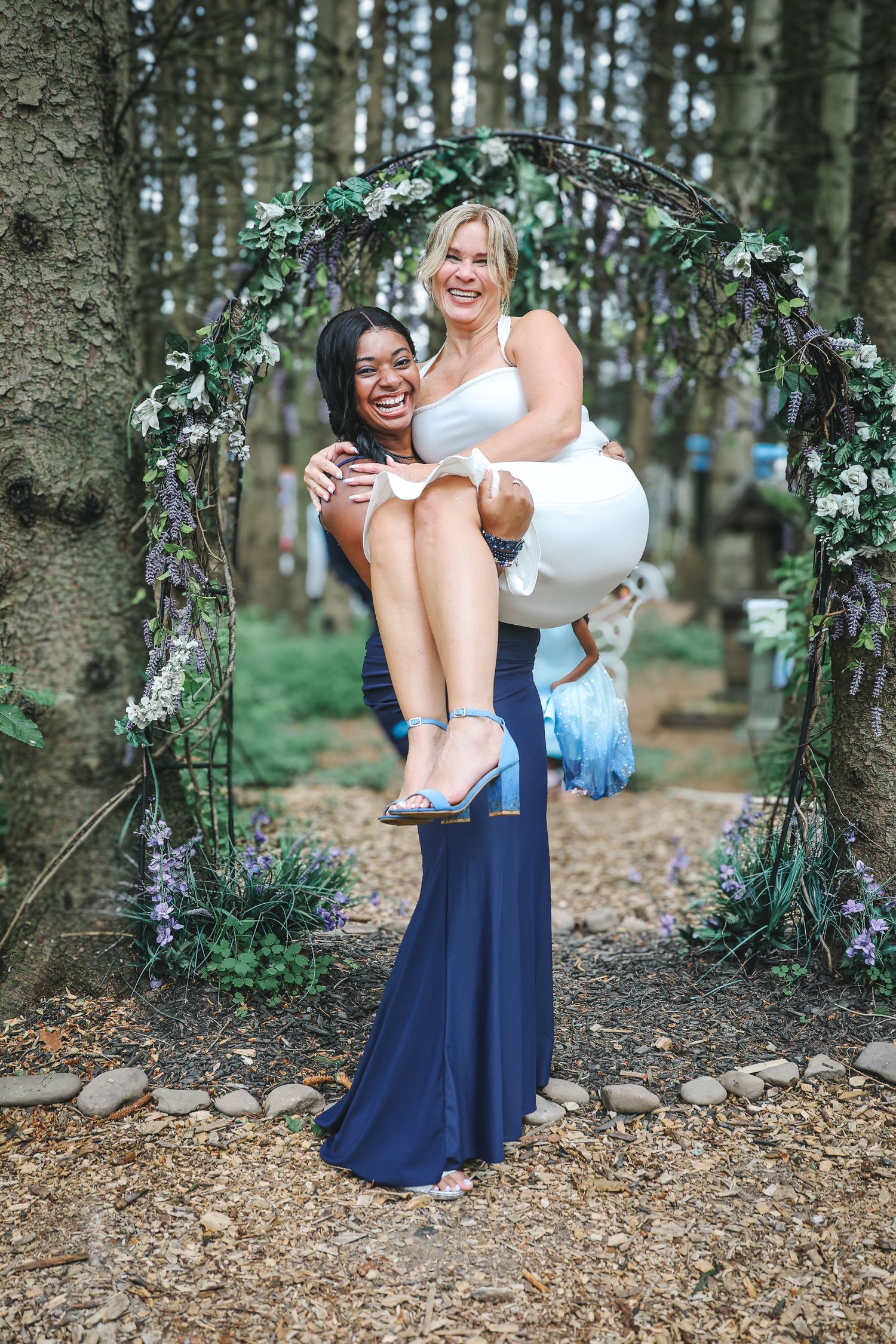 Two friends share a playful moment under a floral arch in a forest setting, one carrying the other in a celebratory pose.