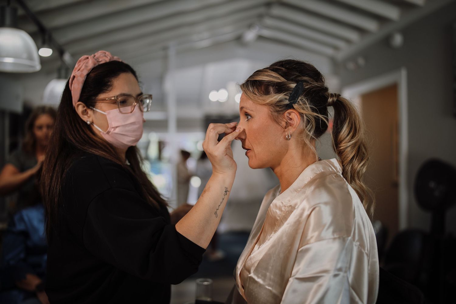 A makeup artist wearing a face mask applies cosmetics to a client in a bright indoor setting.