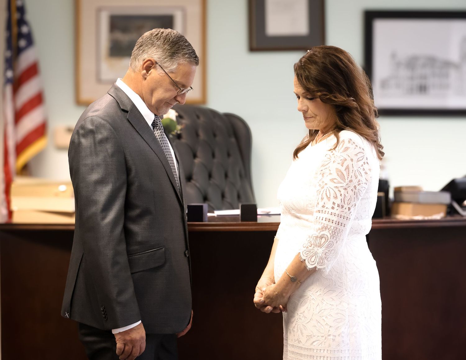 A couple stands together in a courtroom setting with an American flag visible in the background.