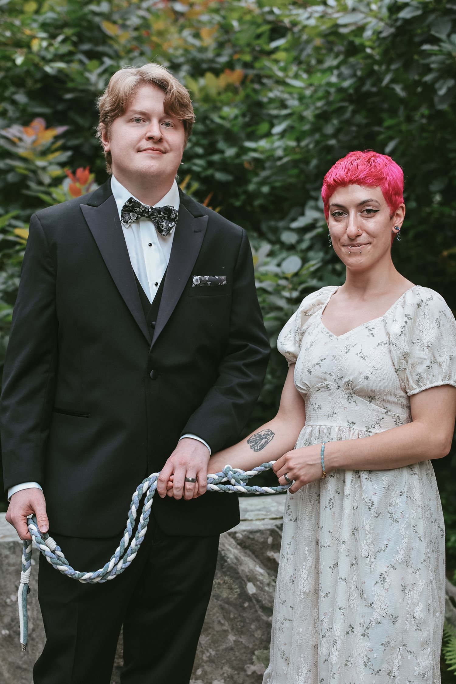 A couple in wedding attire poses together outdoors, with one wearing a black tuxedo and the other in a white dress.