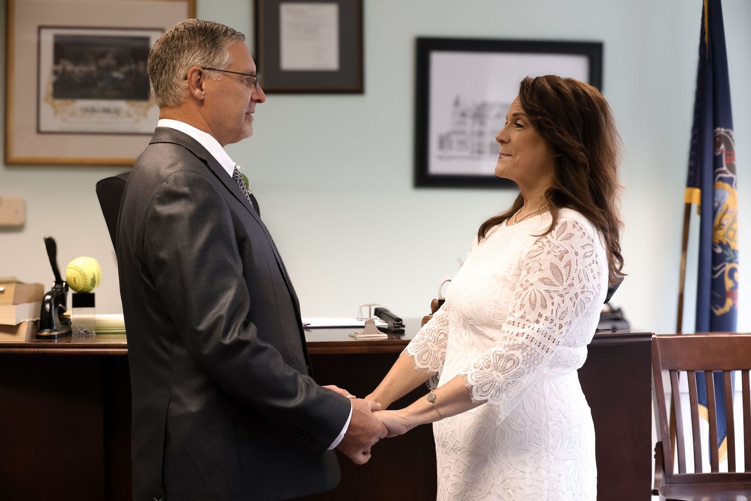 Two people hold hands during an intimate wedding ceremony in an office setting with a desk and flag in the background.