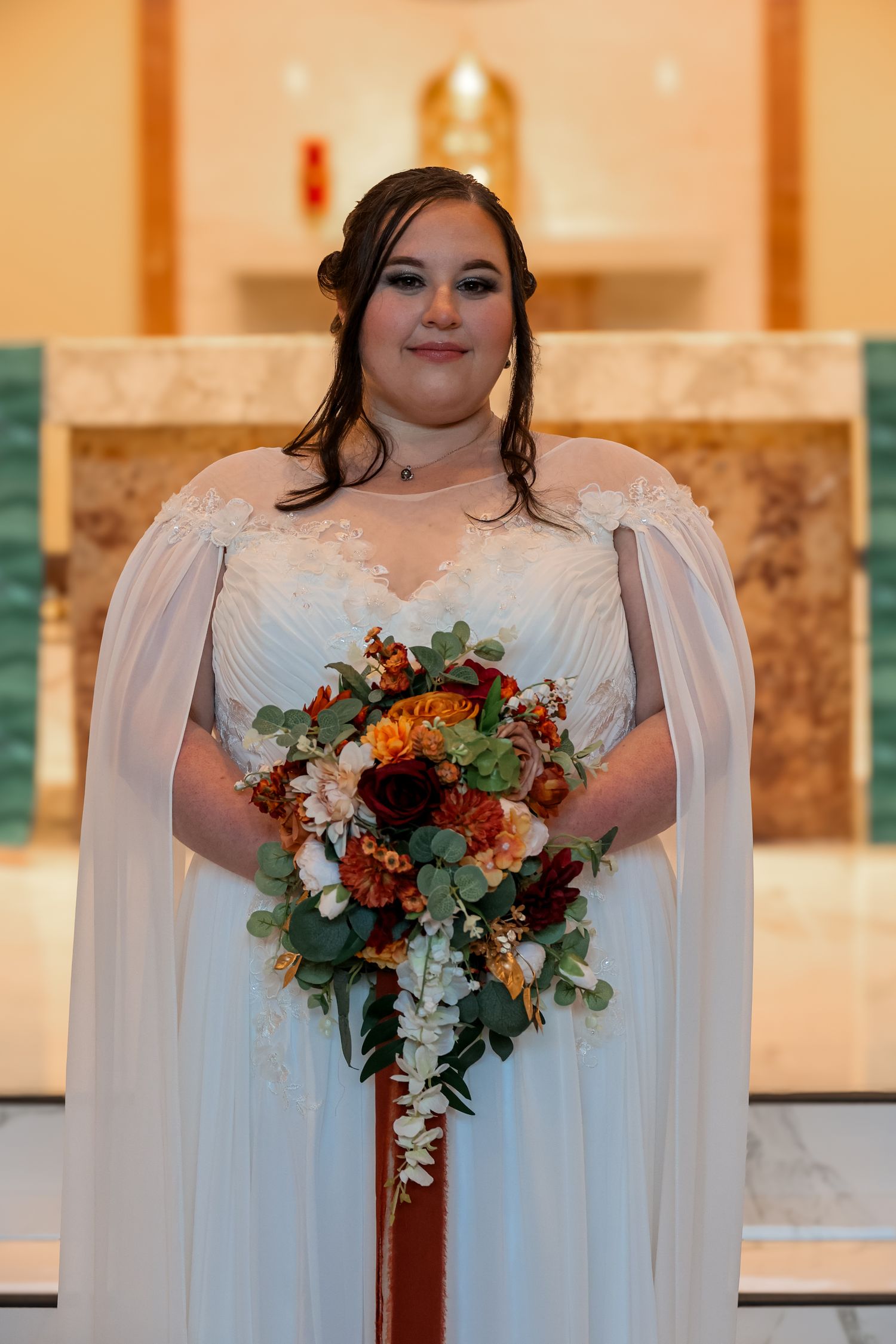 A bride in a flowing white dress holds a vibrant autumn bouquet with burgundy, orange and white flowers and greenery.