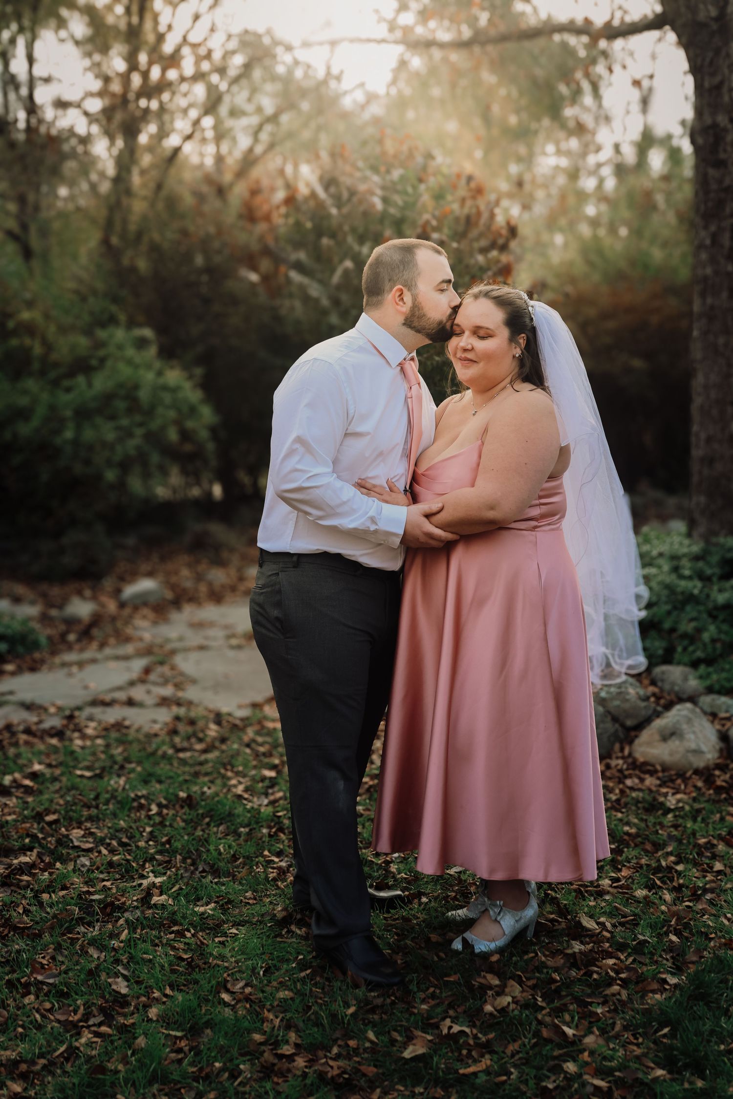 A couple in wedding attire share an intimate moment on fallen autumn leaves in a wooded outdoor setting.
