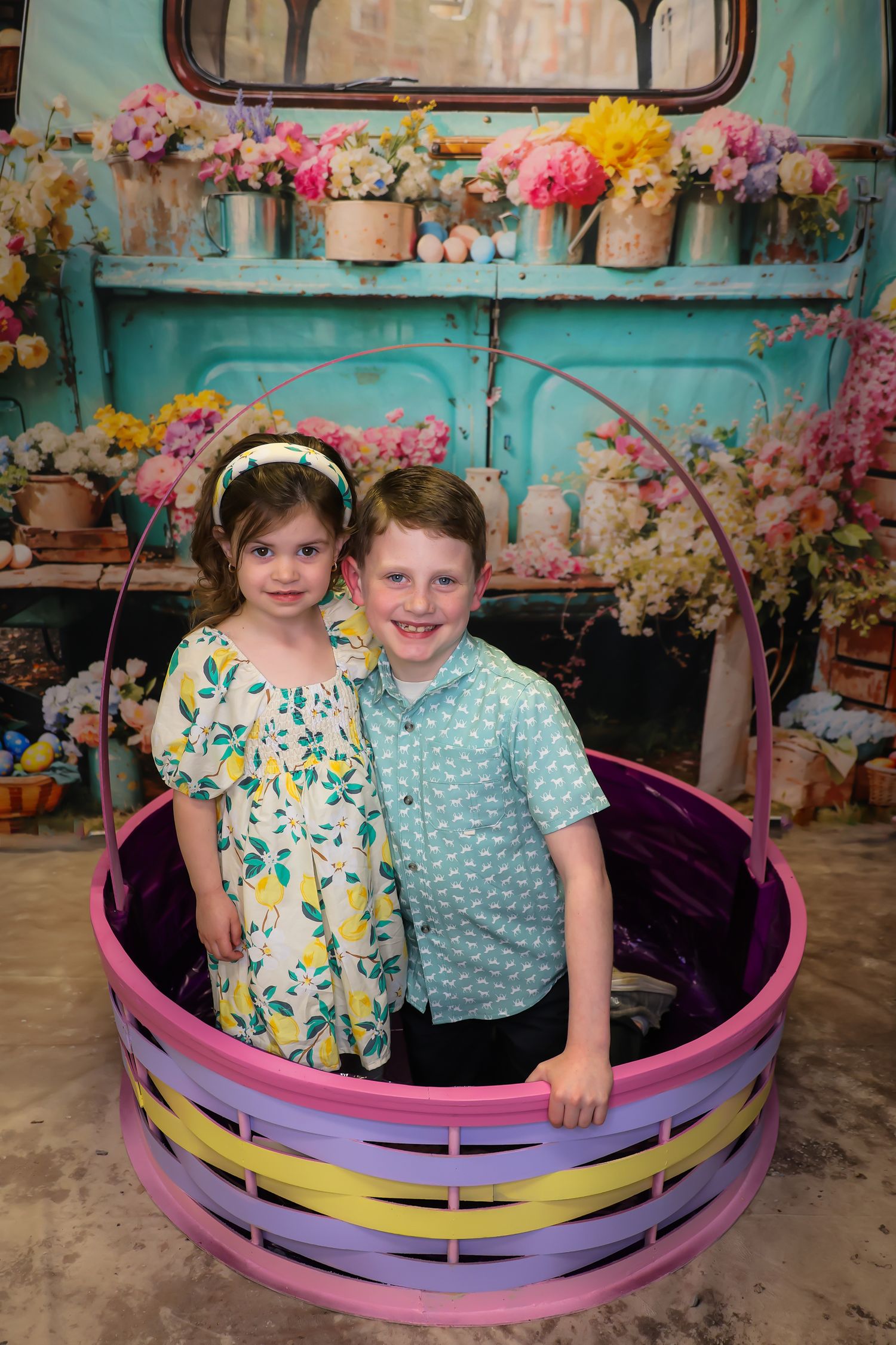 Two young children sit together in a colorful crescent moon prop surrounded by floral decorations and turquoise backdrop.