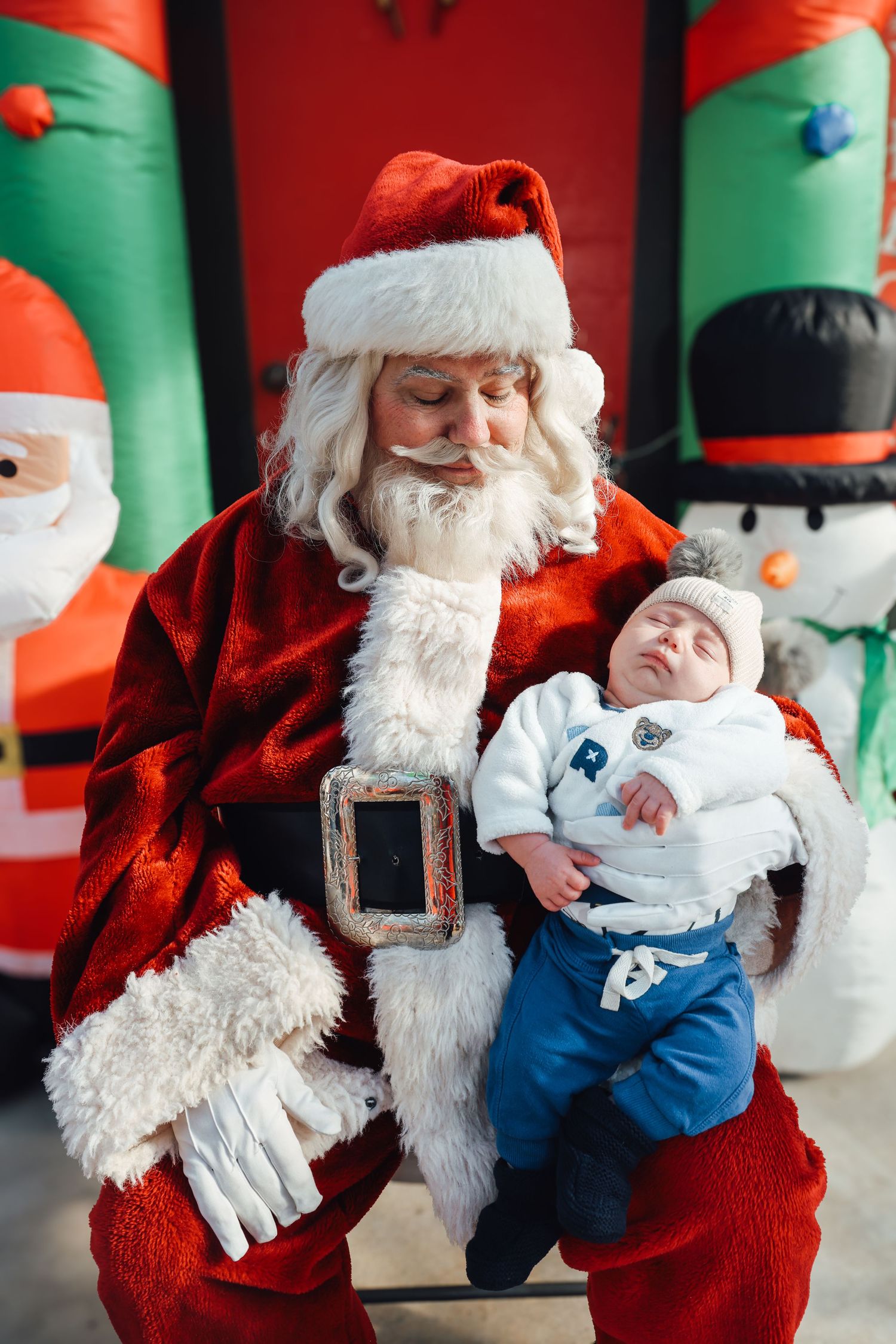 A crying baby sits on the lap of a person in a traditional red and white holiday costume with decorative snowmen in background.