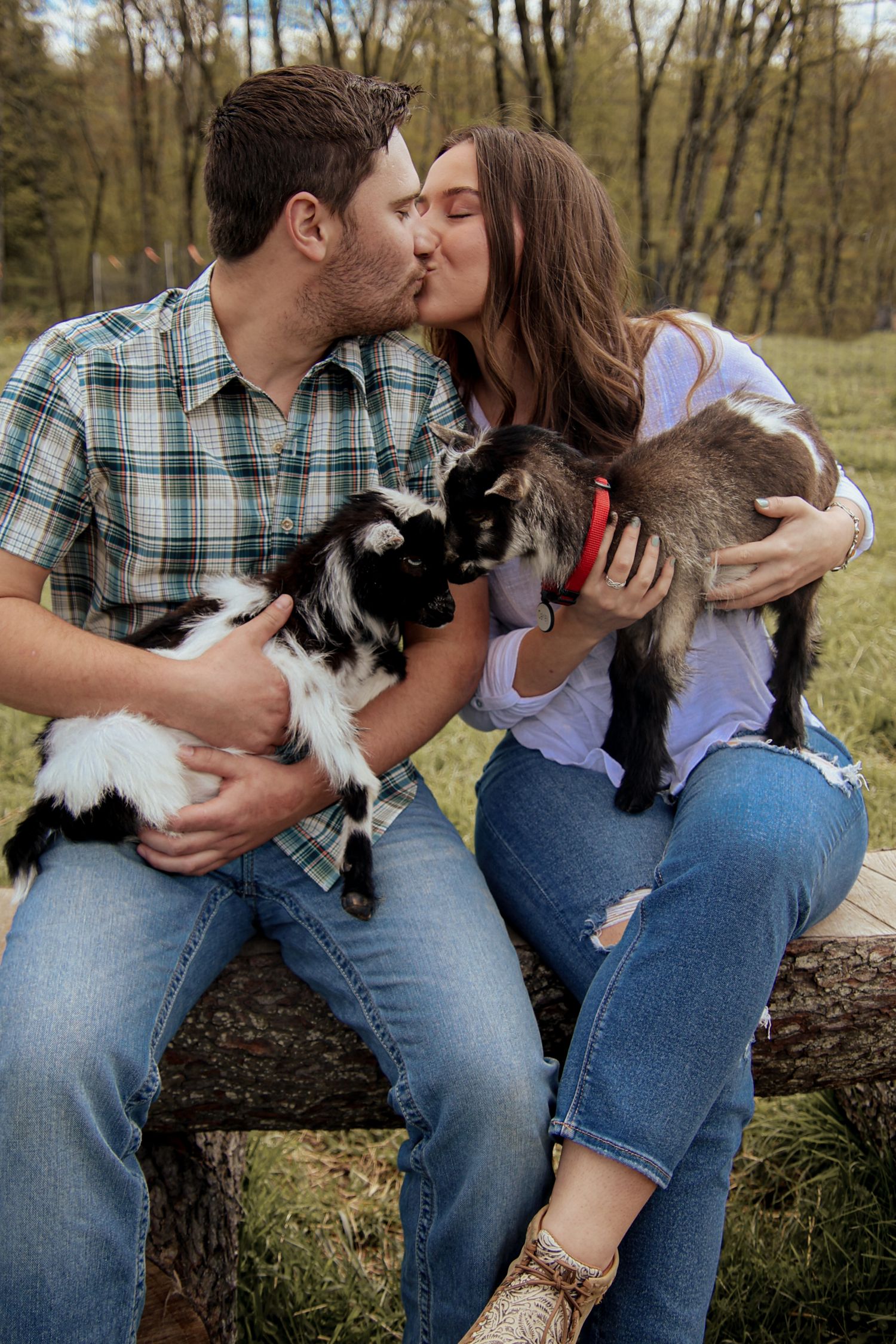 A couple shares a kiss while holding their black and white pets outdoors in a wooded setting.
