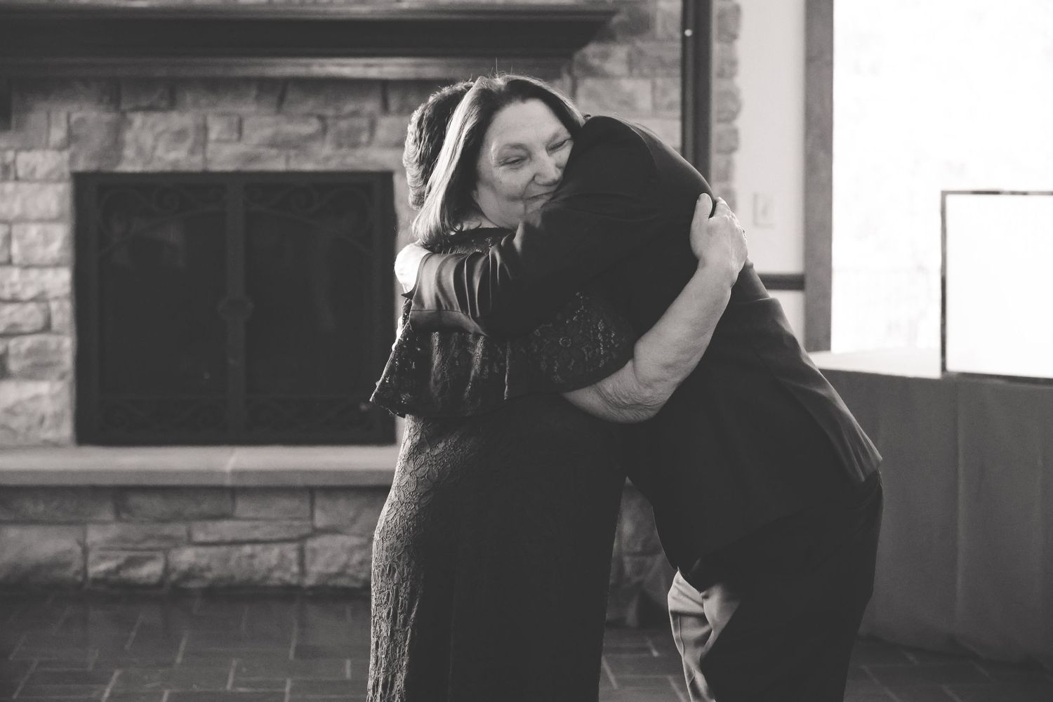A black and white photograph shows two people embracing in front of a brick fireplace.